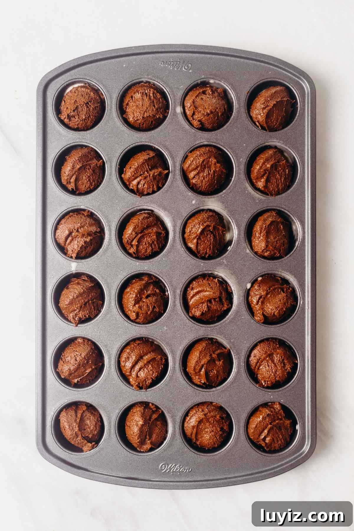 Fudgy Brownie Bites Recipe — Moist Mini Chocolate Brownies 13 Scooped brownie batter in twenty four cavities of a mini muffin tin on a white textured background.