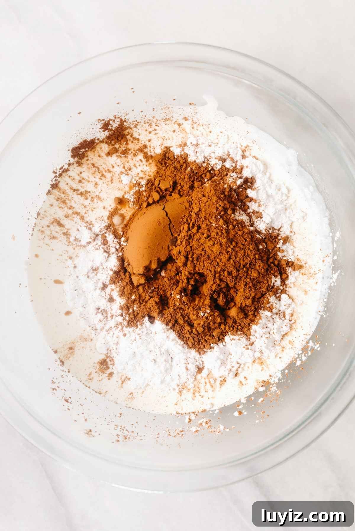 Fudgy Brownie Bites Recipe — Moist Mini Chocolate Brownies 15 Heavy whipping cream, powdered sugar, unsweetened cocoa powder, and vanilla extract added to a chilled mixing bowl on a white textured background.