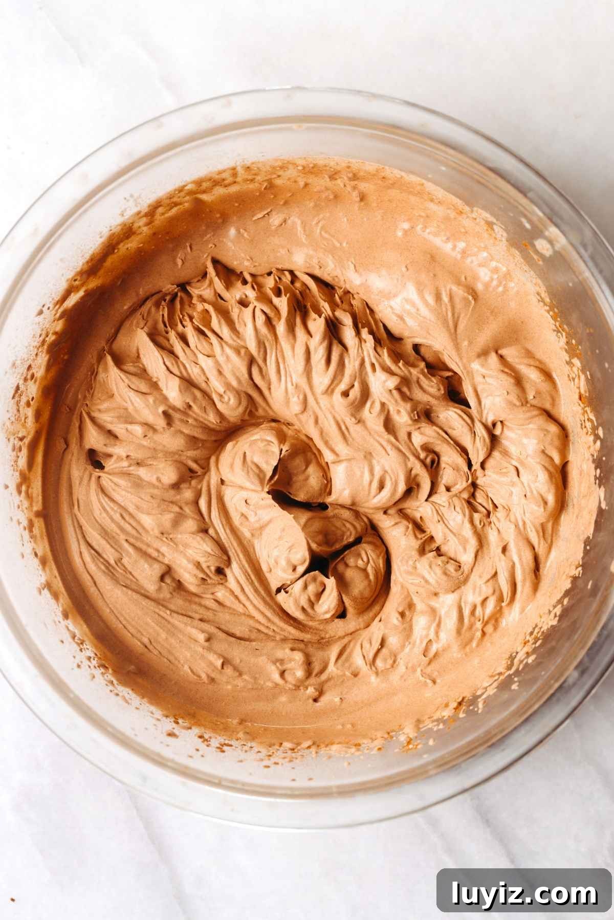 Fudgy Brownie Bites Recipe — Moist Mini Chocolate Brownies 16 Whisked heavy whipping cream, powdered sugar, vanilla extract, and unsweetened cocoa powder in a large chilled mixing bowl on a white textured background.