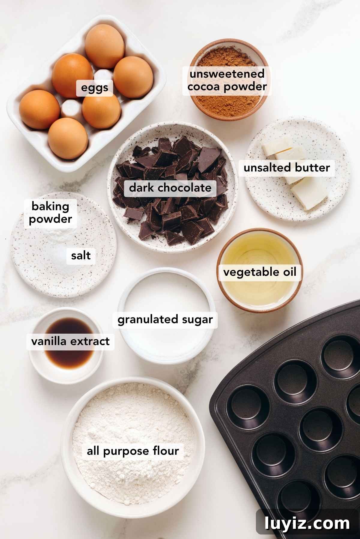 Fudgy Brownie Bites Recipe — Moist Mini Chocolate Brownies 4 Ingredients in various size bowls labeled as eggs, unsweetened cocoa powder, dark chocolate, salt, baking powder, vegetable oil, vanilla extract, granulated sugar, and all purpose flour on a white marbled background.