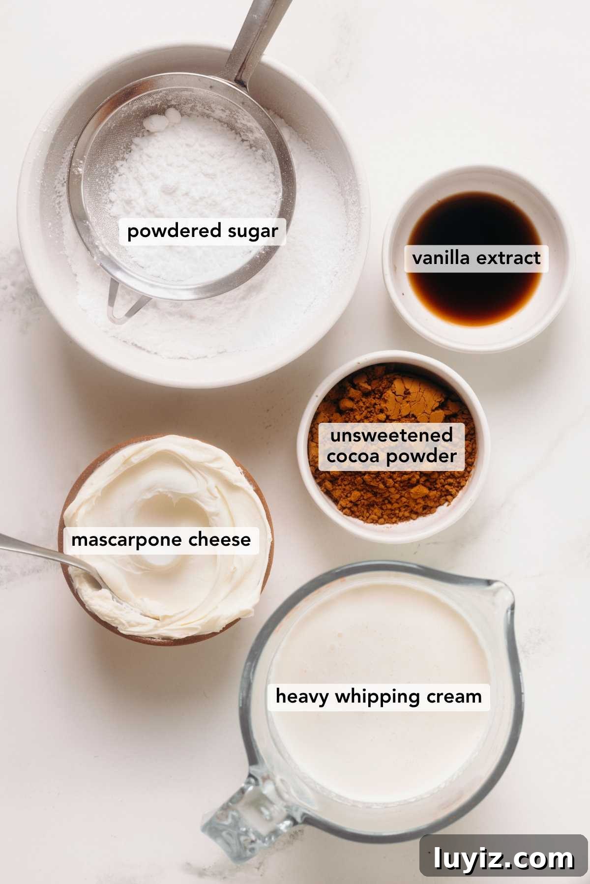 Fudgy Brownie Bites Recipe — Moist Mini Chocolate Brownies 5 Ingredients in various size bowls labeled as powdered sugar, vanilla extract, unsweetened cocoa powder, mascarpone cheese, and heavy whipping cream on a white marbled background.