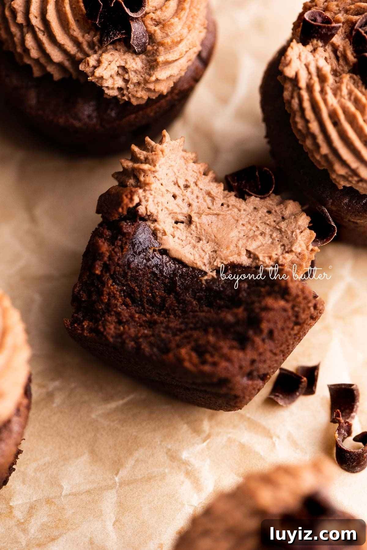 Fudgy Brownie Bites Recipe — Moist Mini Chocolate Brownies 6 Brownie bites topped with chocolate mascarpone whipped cream and chocolate curls on a wrinkled brown parchment paper background with bite taken out of center brownie.