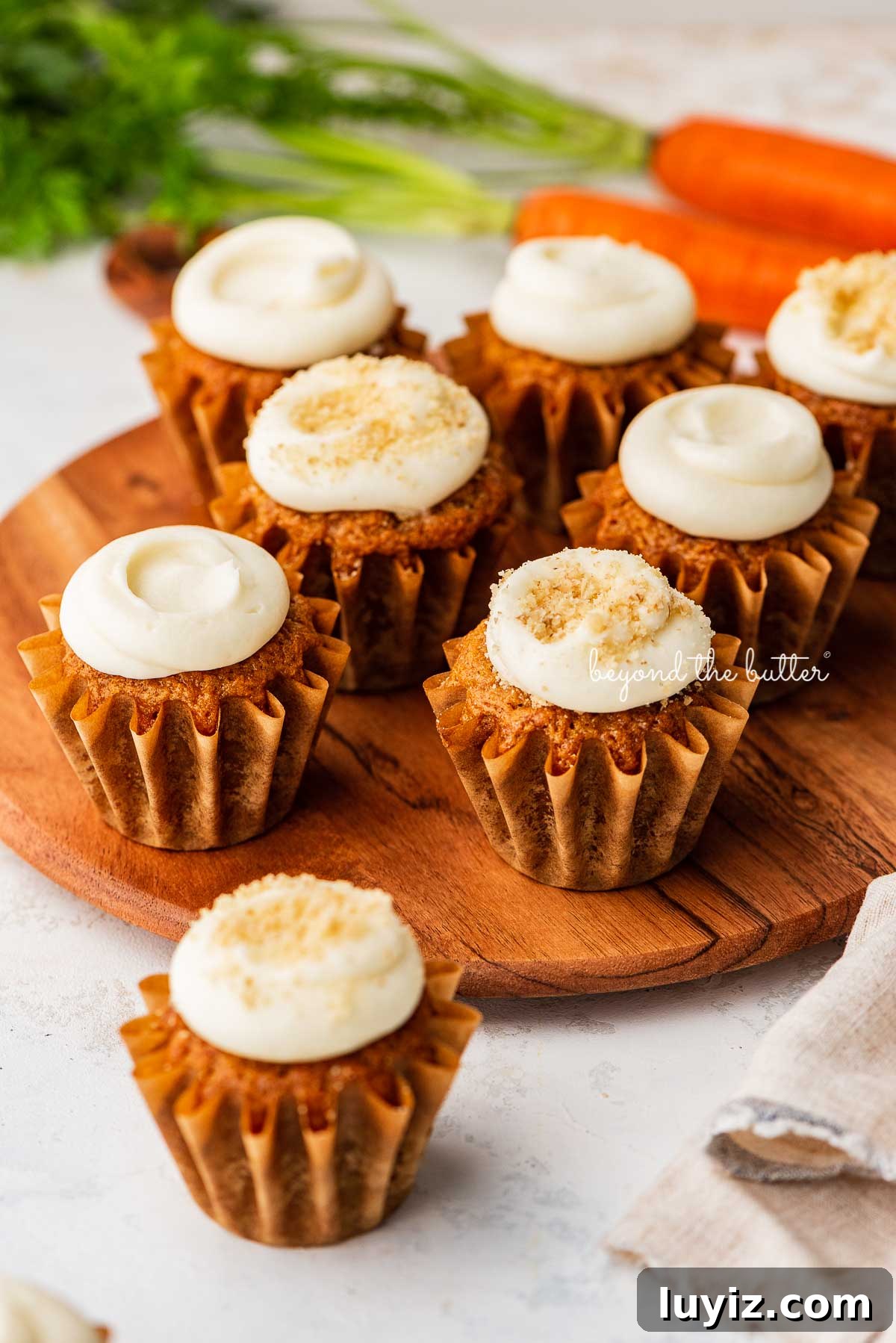 Ultra-Moist Carrot Cake Cupcakes with Cream Cheese Frosting 3 Super moist carrot cake cupcakes with cream cheese frosting placed on a wood cake stand on a white background with some carrots | © Beyond the Butter®
