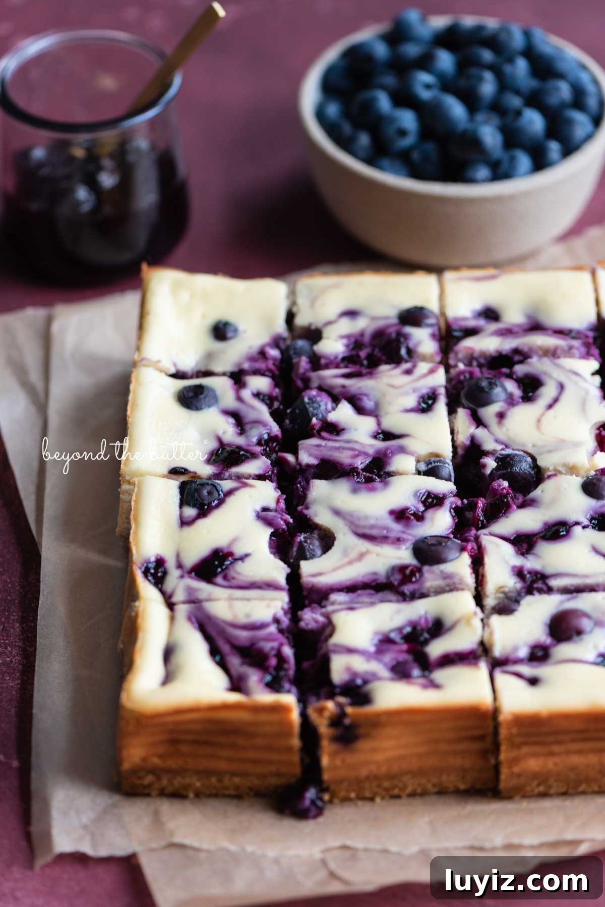 Blueberry Swirl Cheesecake Bars Recipe — Creamy Layered Dessert 3 Slices of blueberry swirl cheesecake bars placed on two sheets of parchment paper with bowl of blueberry sauce and fresh blueberries to the side on pink background.