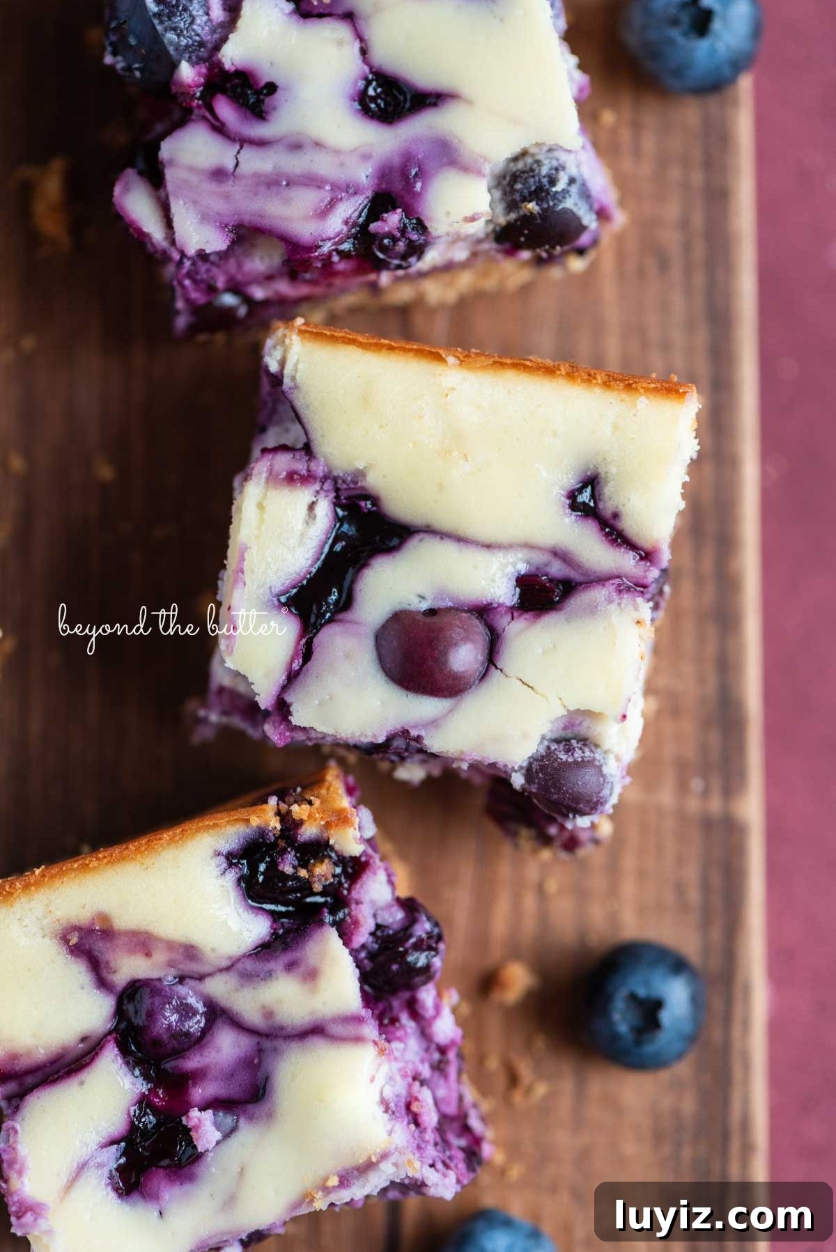 Blueberry Swirl Cheesecake Bars Recipe — Creamy Layered Dessert 9 Sliced blueberry swirl cheesecake bars randomly placed on a wood cutting board with blueberries on a pink background.