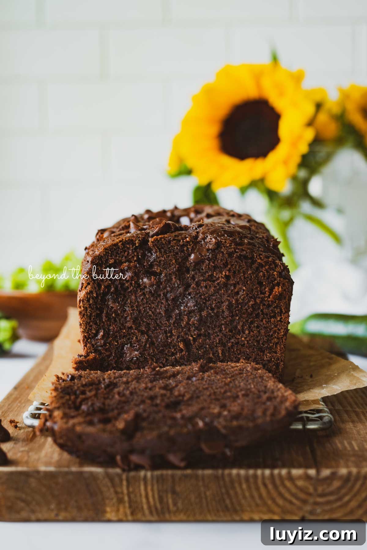 One-Bowl Chocolate Zucchini Quick Bread Recipe 6 Sliced chocolate zucchini bread on a cutting board with white tile background and sunflowers | © Beyond the Butter®