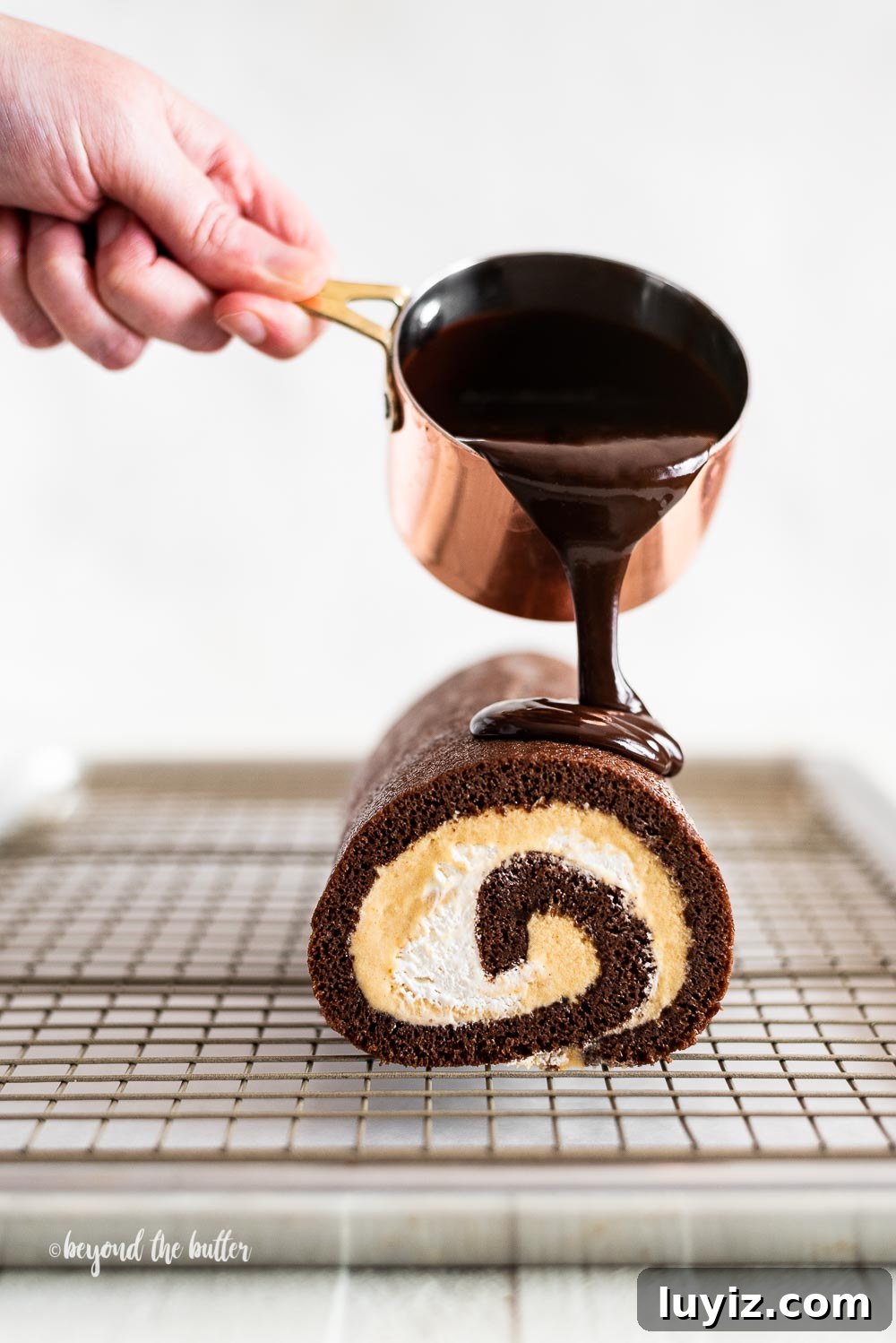 Chocolate Pumpkin Swiss Roll with Cocoa Cream Filling 2 Pouring melted chocolate from small copper pot over chocolate pumpkin roll on wire cooling rack.
