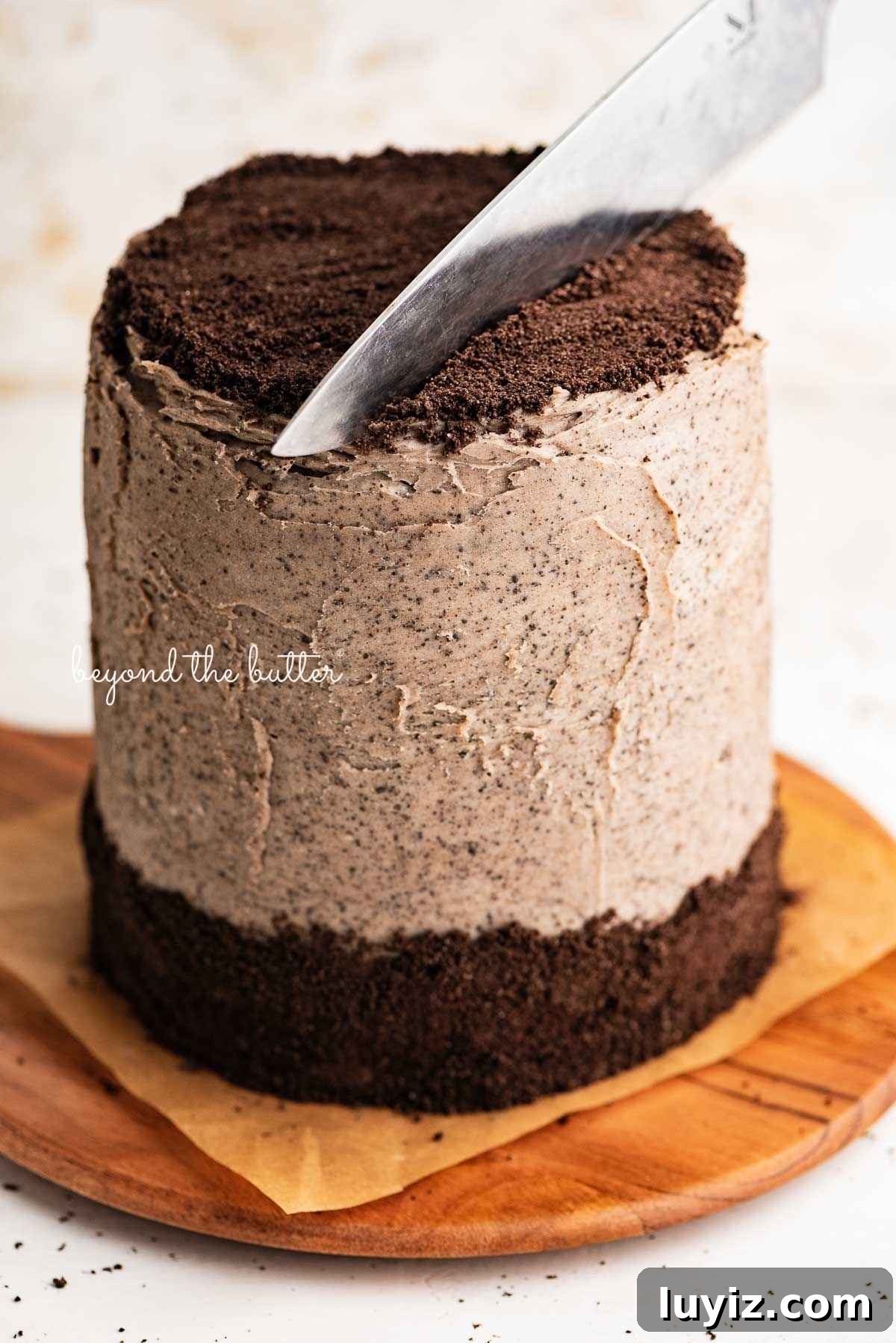 Decadent Chocolate Cake with Oreo Buttercream Frosting 18 Big knife cutting into a three layer chocolate cake with oreo buttercream frosting on a parchment paper covered wood cake board on a white and beige painted background.