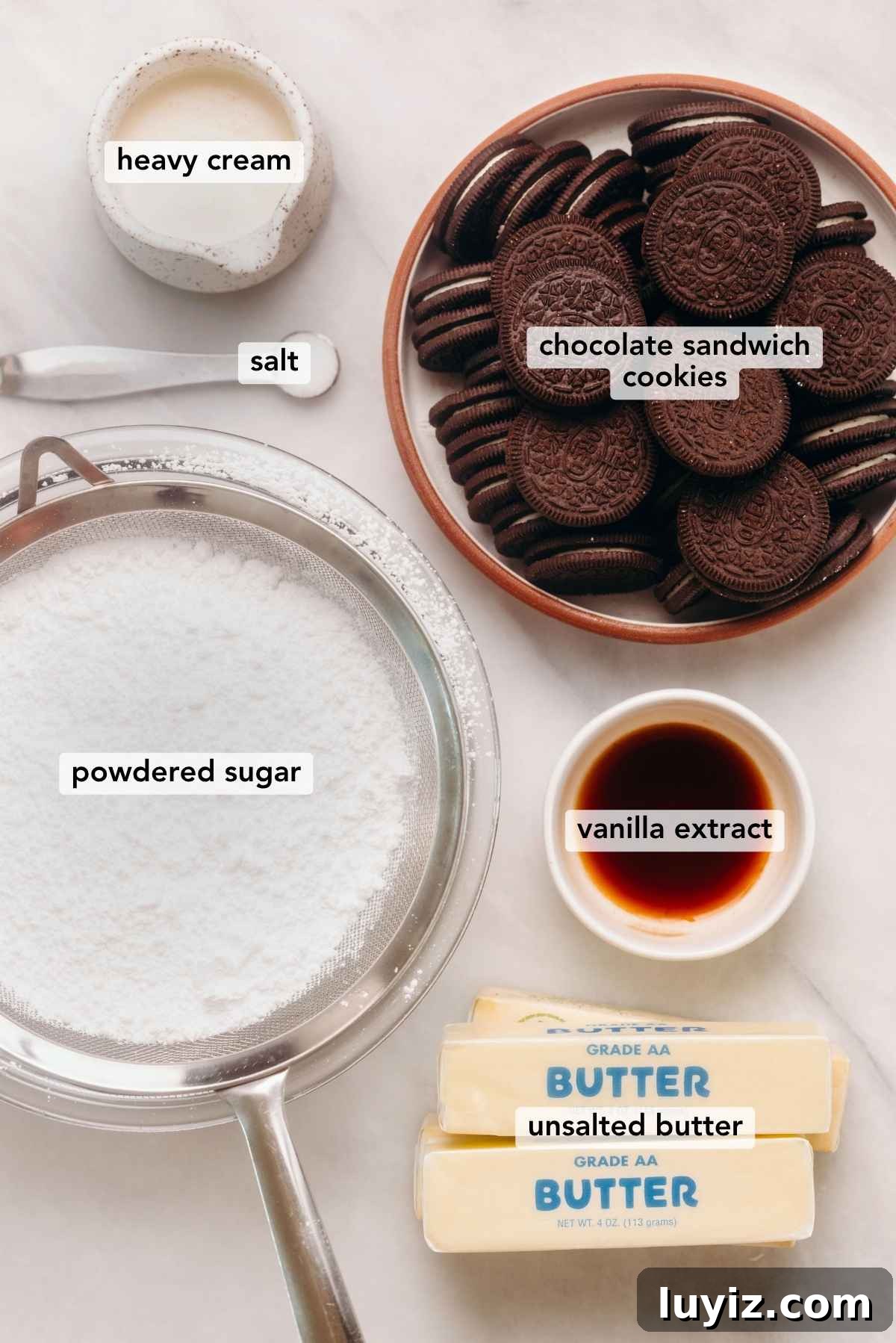 Decadent Chocolate Cake with Oreo Buttercream Frosting 5 Ingredients in various size bowls labeled as heavy cream, chocolate sandwich cookies, salt, powdered sugar, vanilla extract, and unsalted butter on a white marbled background.