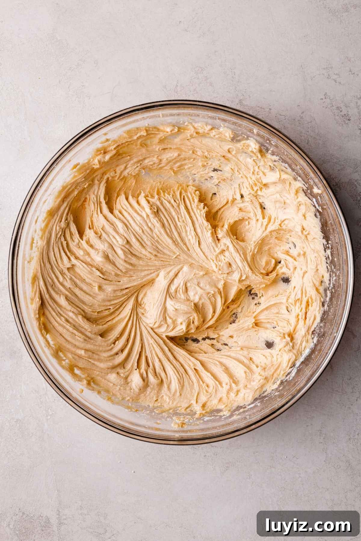 Butterscotch Krimpet Sheet Cake Recipe — Moist Retro Dessert 14 Butterscotch frosting whisked in a large glass bowl on a white and grey textured background.