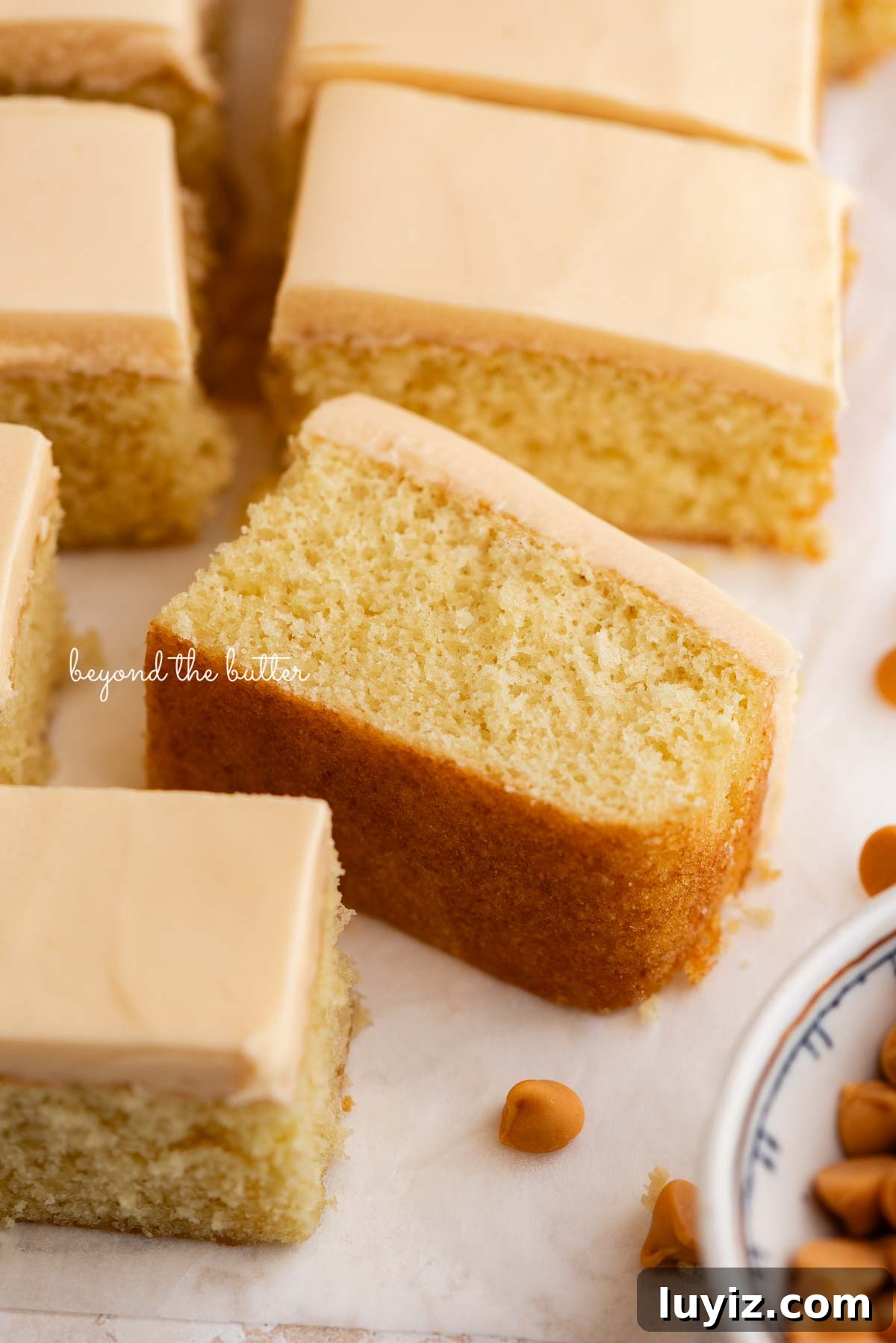 Butterscotch Krimpet Sheet Cake Recipe — Moist Retro Dessert 4 Slices of butterscotch krimpets placed on a parchment paper lined background with one slice on its side with a small decorative ceramic bowl of butterscotch chips placed next to the cake.