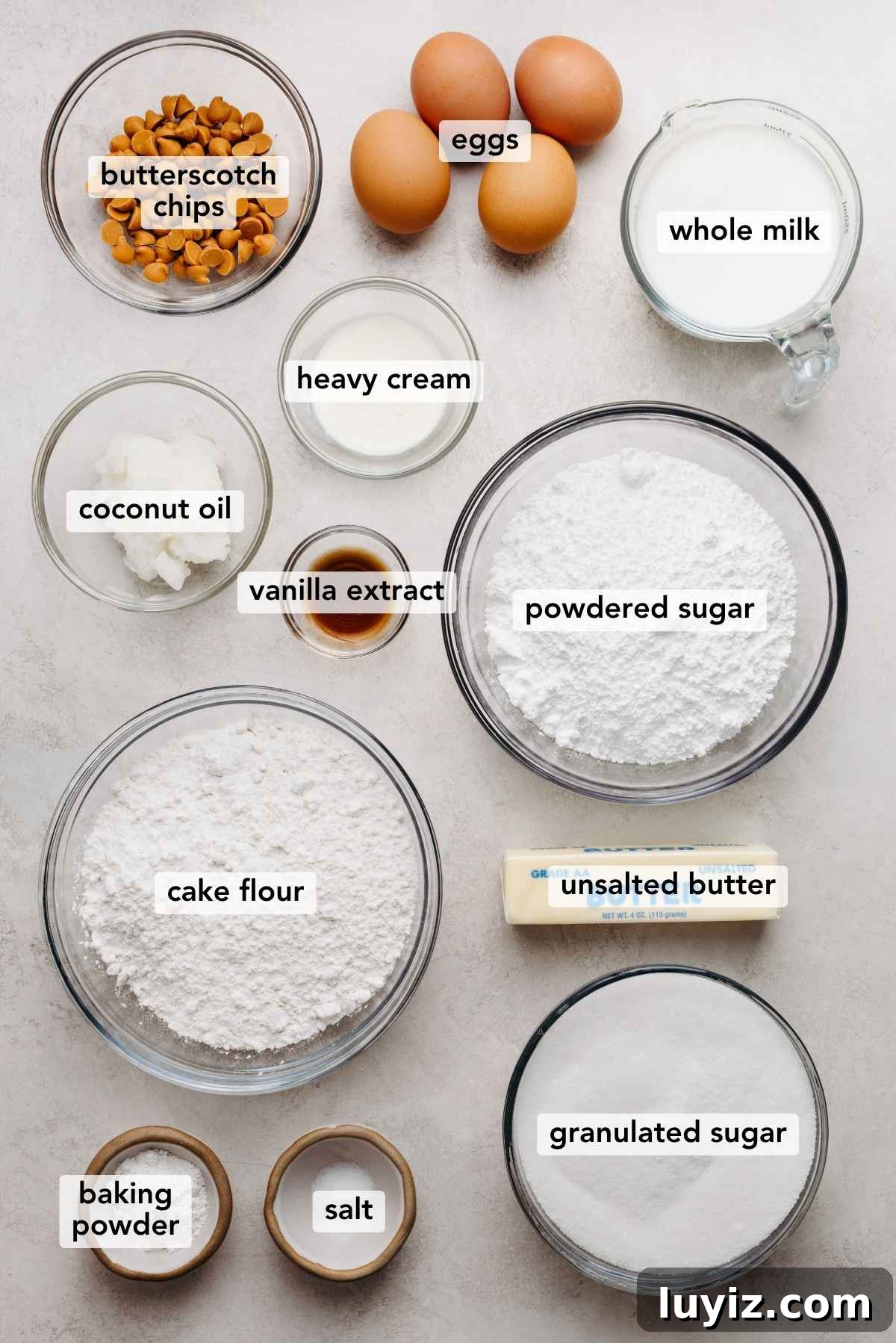 Butterscotch Krimpet Sheet Cake Recipe — Moist Retro Dessert 5 Ingredients in various size bowls labeled as butterscotch chips, eggs, whole milk, coconut oil, heavy cream, vanilla extract, powdered sugar, cake flour, unsalted butter, baking powder, salt, and granulated sugar on a white marbled background.