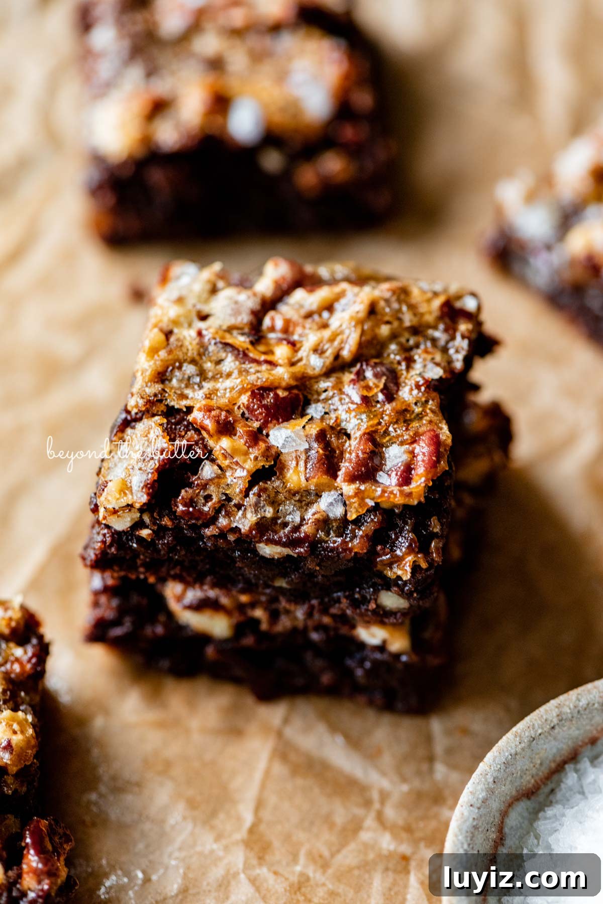 Salted Pecan Pie Brownies Recipe: Gooey Chocolate with Crunch 6 Stacked salted pecan pie brownies on brown parchment paper | © Beyond the Butter®