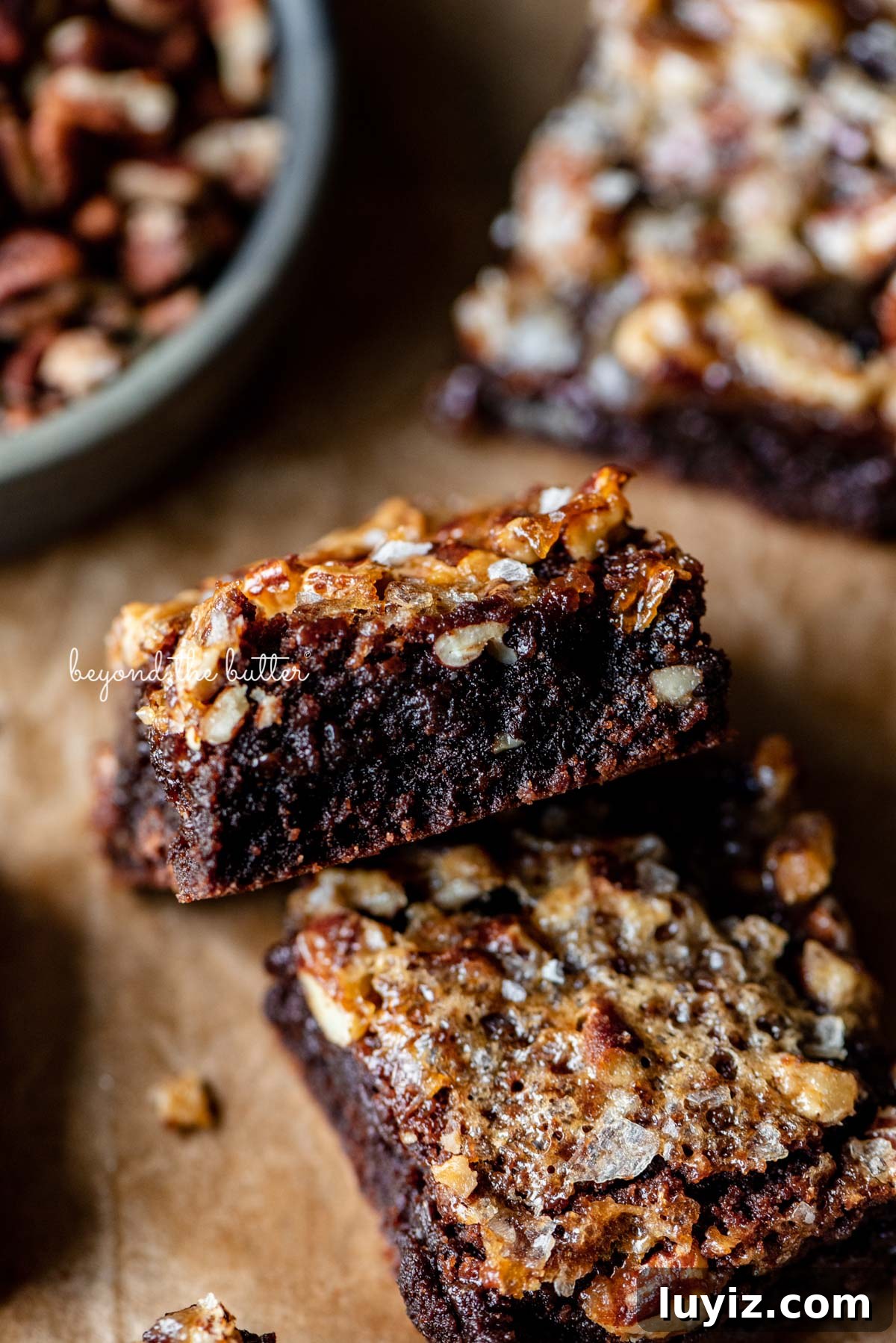 Salted Pecan Pie Brownies Recipe: Gooey Chocolate with Crunch 8 Semi-stacked salted pecan pie brownies on brown parchment paper | © Beyond the Butter®
