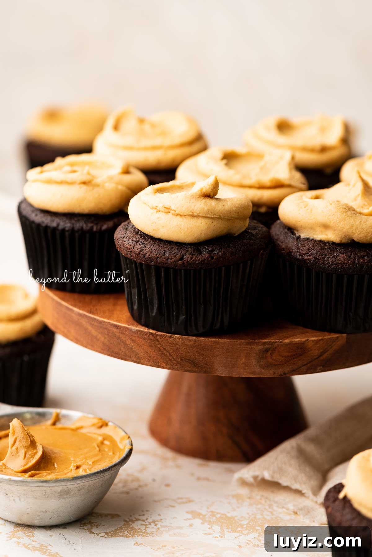 Decadent Chocolate Cupcakes with Peanut Butter Frosting 3 Wood cake stand with devil's food cupcakes surrounded by other cupcakes, peanut butter, and a napkin on a white background | © Beyond the Butter®