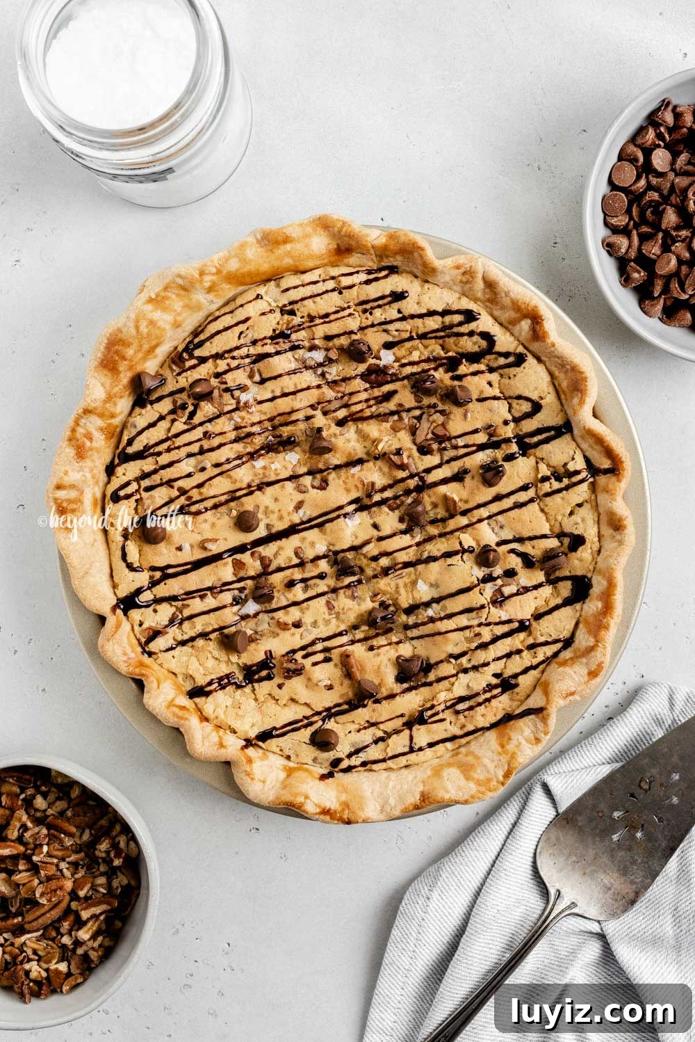 Ultimate Chocolate Chip Pie Recipe: Gooey, Crispy, and Irresistible 3 Overhead image of chocolate chip pie drizzled with chocolate syrup with bowls of chocolate chips, pecans, and a server