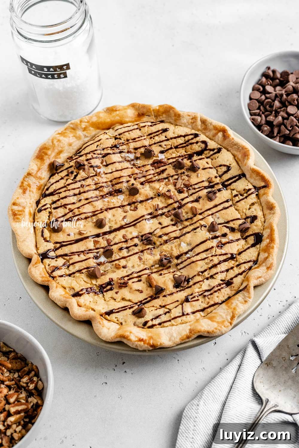 Ultimate Chocolate Chip Pie Recipe: Gooey, Crispy, and Irresistible 8 Overhead angled image of chocolate chip pie drizzled with chocolate syrup with bowls of chocolate chips, pecans, and a server