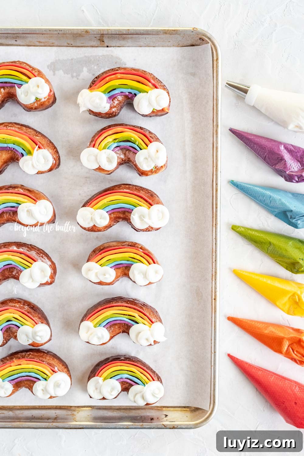 How to Make Vibrant Rainbow Donuts at Home 8 Overhead image of just decorated rainbow donuts on a baking sheet with piping bags filled with buttercream | All Images © Beyond the Butter, LLC