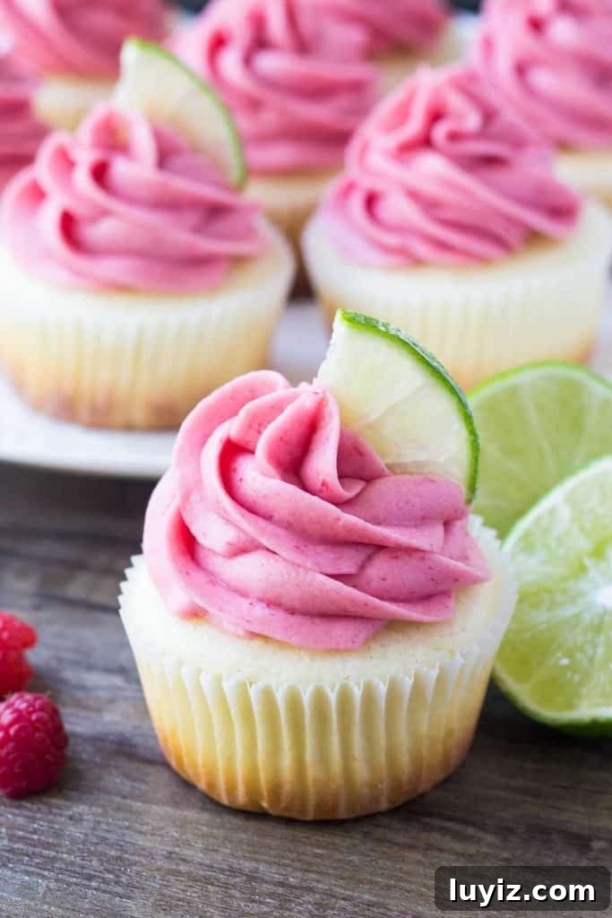 Lime Cupcakes with Raspberry Buttercream Frosting 3 One lime cupcake with raspberry frosting and a plate of cupcakes in the background. Garnished with lime slices and raspberries.