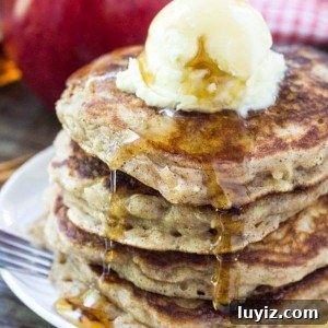 Stack of Apple Pancakes with Brown Sugar and Cinnamon. Topped with whipped butter and drizzled with maple syrup. Apple and bottle of syrup in background.