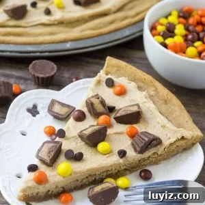 Slice of peanut butter cookie pizza topped with peanut butter frosting, peanut butter cups and peanut butter candies. Bowl of Reese's Pieces and whole cookie pizza in the background.