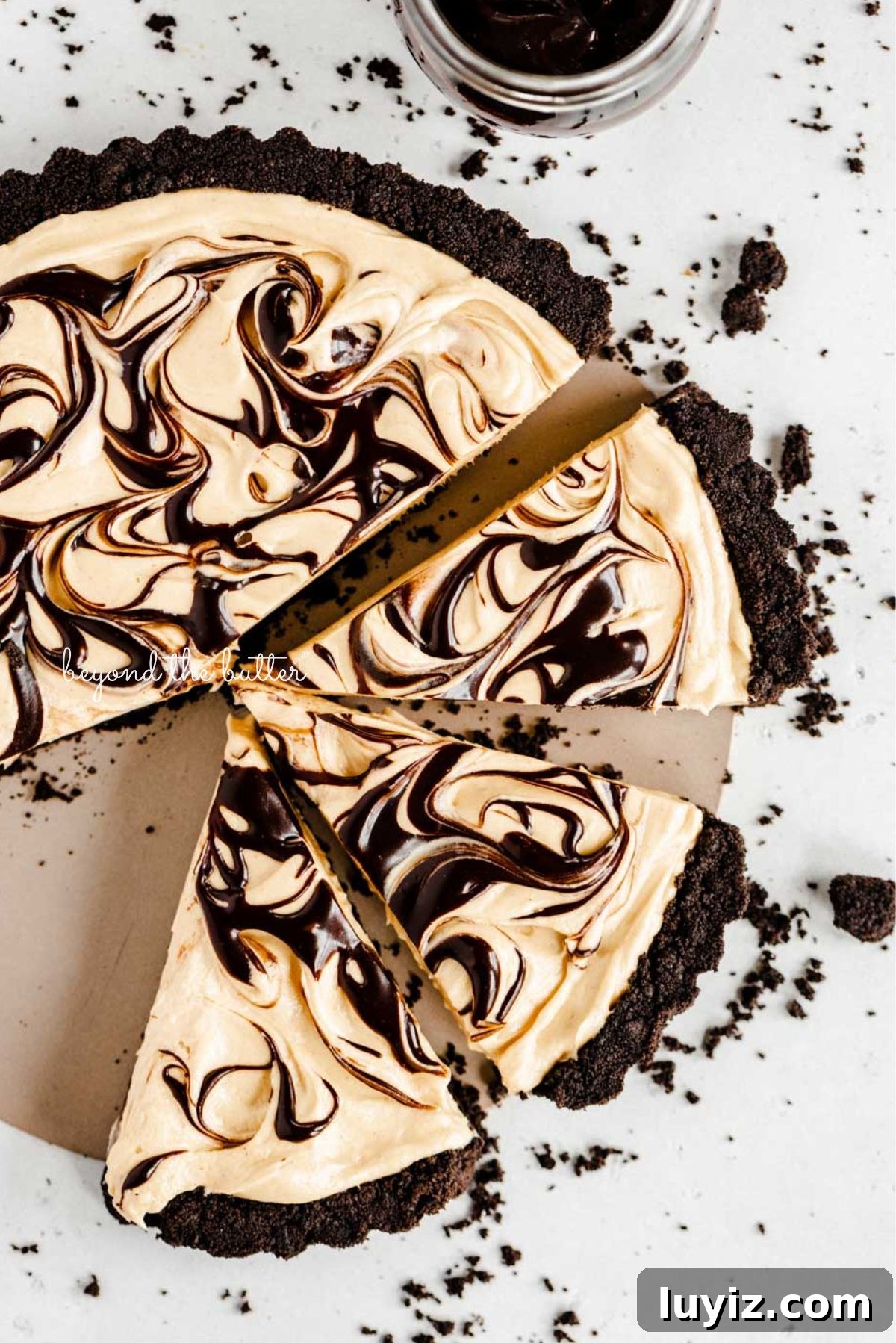 Chocolate Peanut Butter Swirl Tart Recipe — Decadent No-Bake Dessert 2 Round chocolate peanut butter tart swirled with chocolate syrup and cut into slices with a bowl of chocolate sandwich crumbs next to it on a white speckled background.