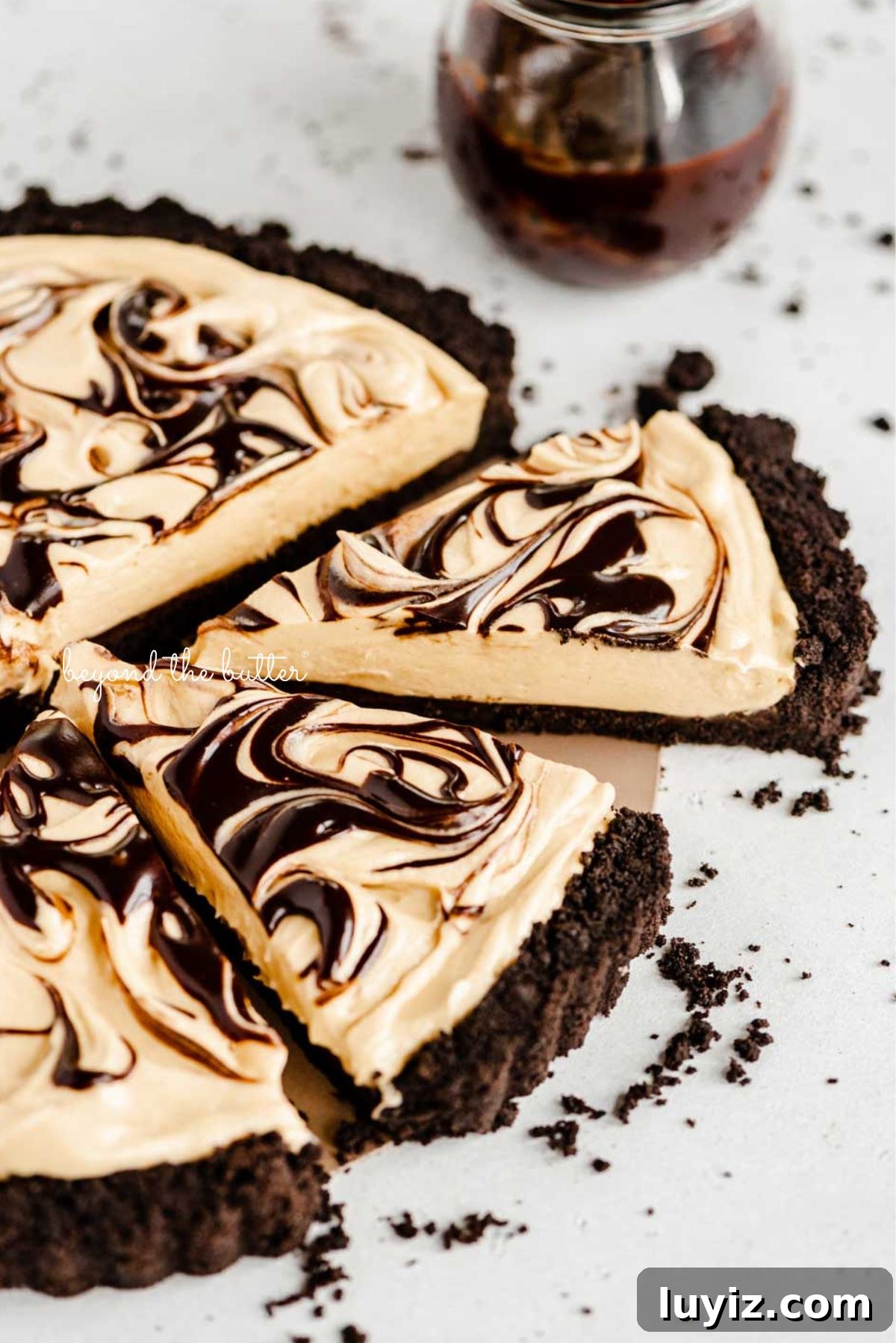 Chocolate Peanut Butter Swirl Tart Recipe — Decadent No-Bake Dessert 3 Round chocolate peanut butter tart swirled with chocolate syrup and cut into slices with a bowl of chocolate syrup next to it surrounded by a few chocolate sandwich cookie crumbs on a white speckled background.