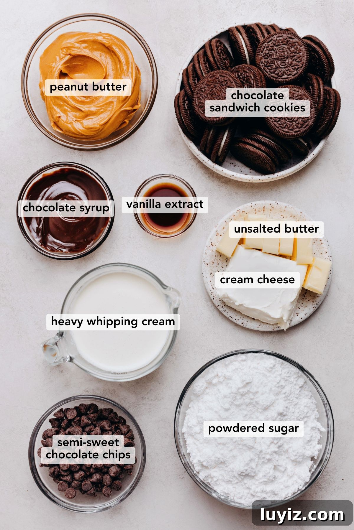 Chocolate Peanut Butter Swirl Tart Recipe — Decadent No-Bake Dessert 4 Ingredients in various size bowls labeled as peanut butter, chocolate sandwich cookies, chocolate syrup, vanilla extract, unsalted butter, cream cheese, heavy whipping cream, semi-sweet chocolate chips, and powdered sugar on a white marbled background.