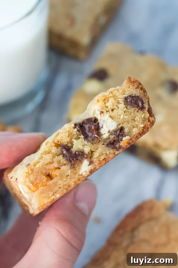 Triple-Chip Cookie Bar Recipe: Chewy Chocolate, Peanut & Butterscotch 3 Side shot of a hand holding a triple chip cookie bar. Glass of milk and more cookie bars in the background.