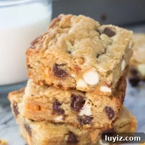 Side shot of 3 stacked triple chip cookie bars with a glass of milk and the cookie bar pan in the background.