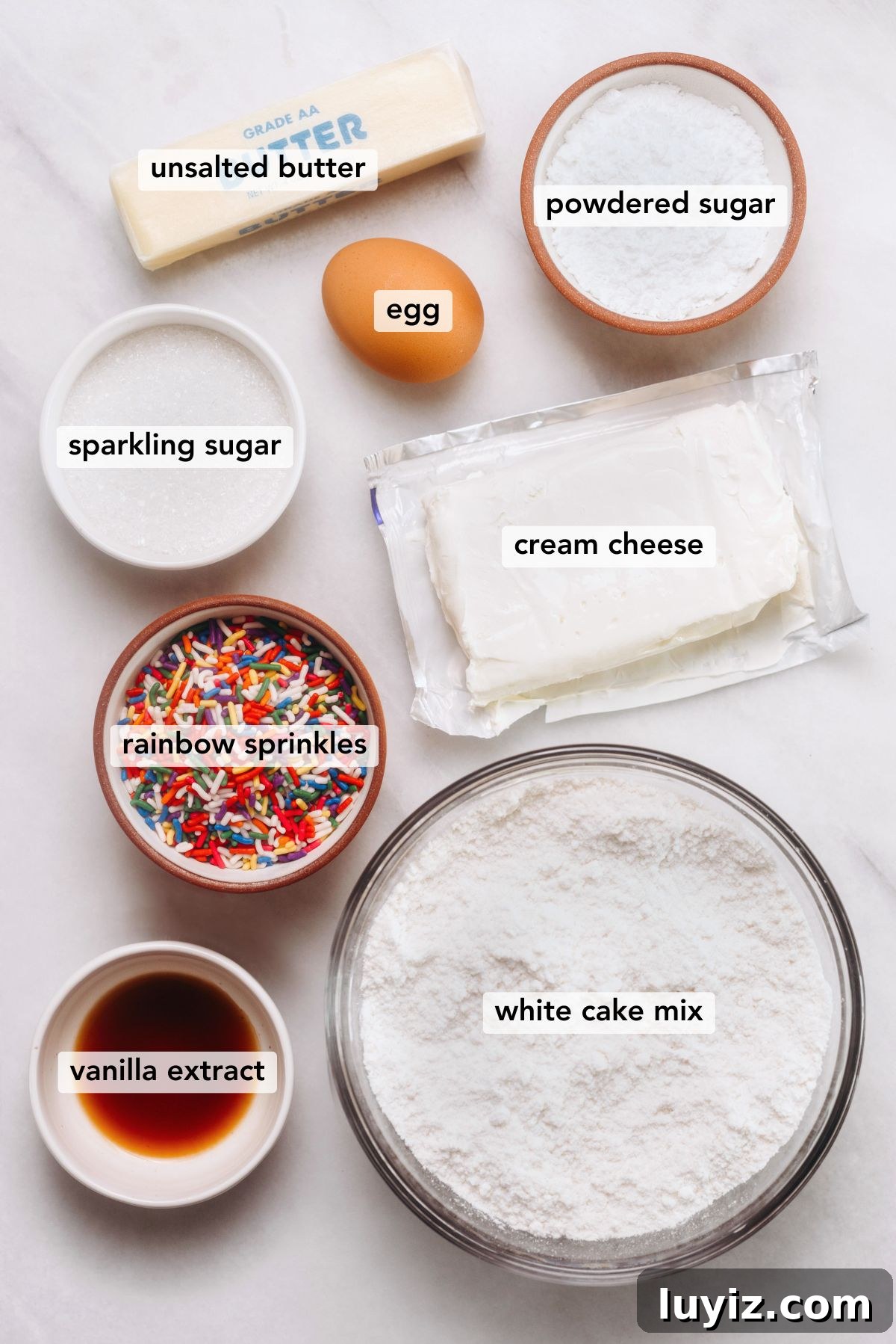 Funfetti Butter Cookies Recipe with Sprinkle-Filled Shortbread 4 Ingredients in various size bowls labeled as all unsalted butter, powdered sugar, egg, sparkling sugar, cream cheese, rainbow sprinkles, white cake mix, and vanilla extract on a white marbled background.