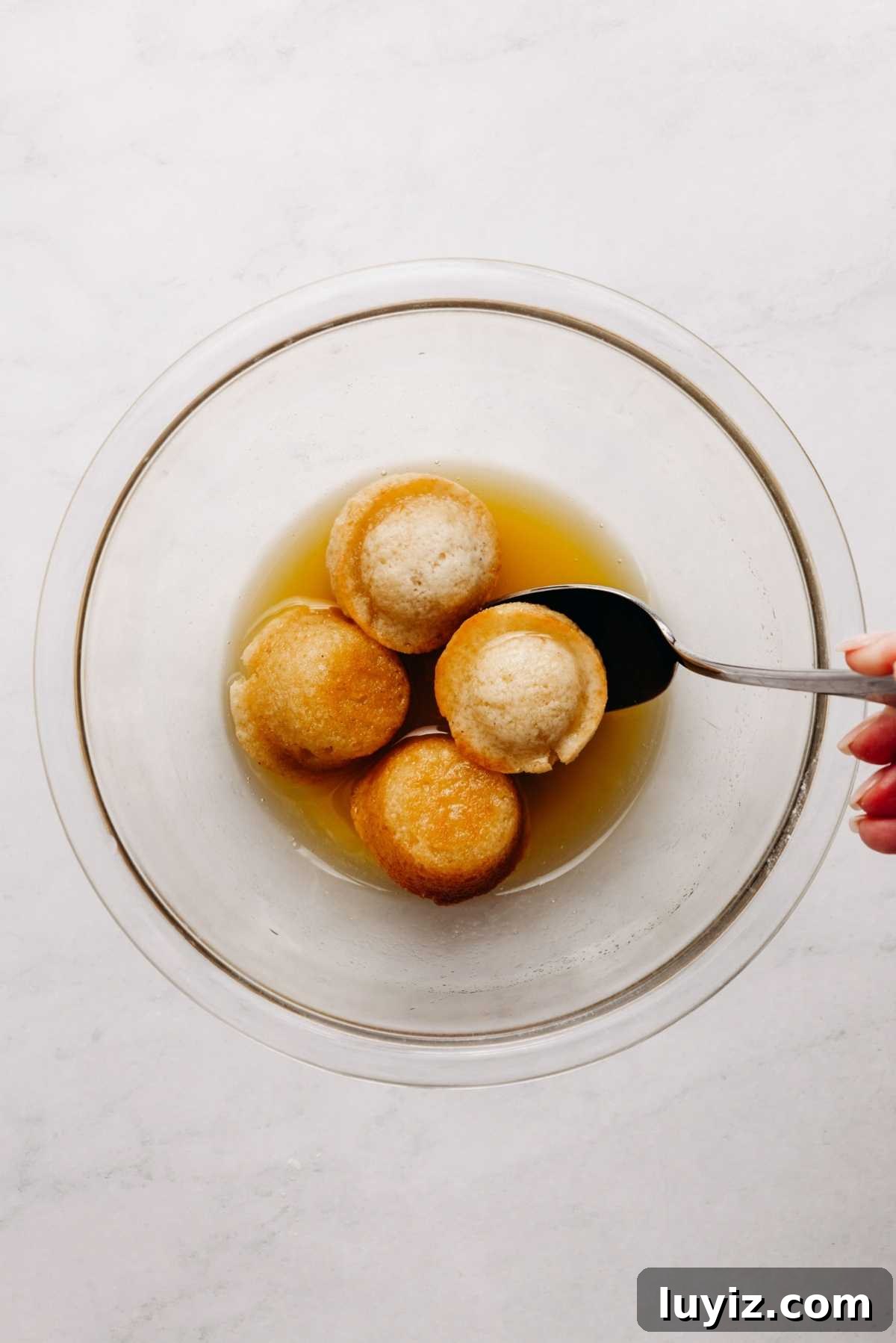 Mini French Breakfast Puffs: Airy, Jam-Filled Pastry Bites 12 Coating mini breakfast muffins in melted butter in a small glass bowl on a white marbled background.