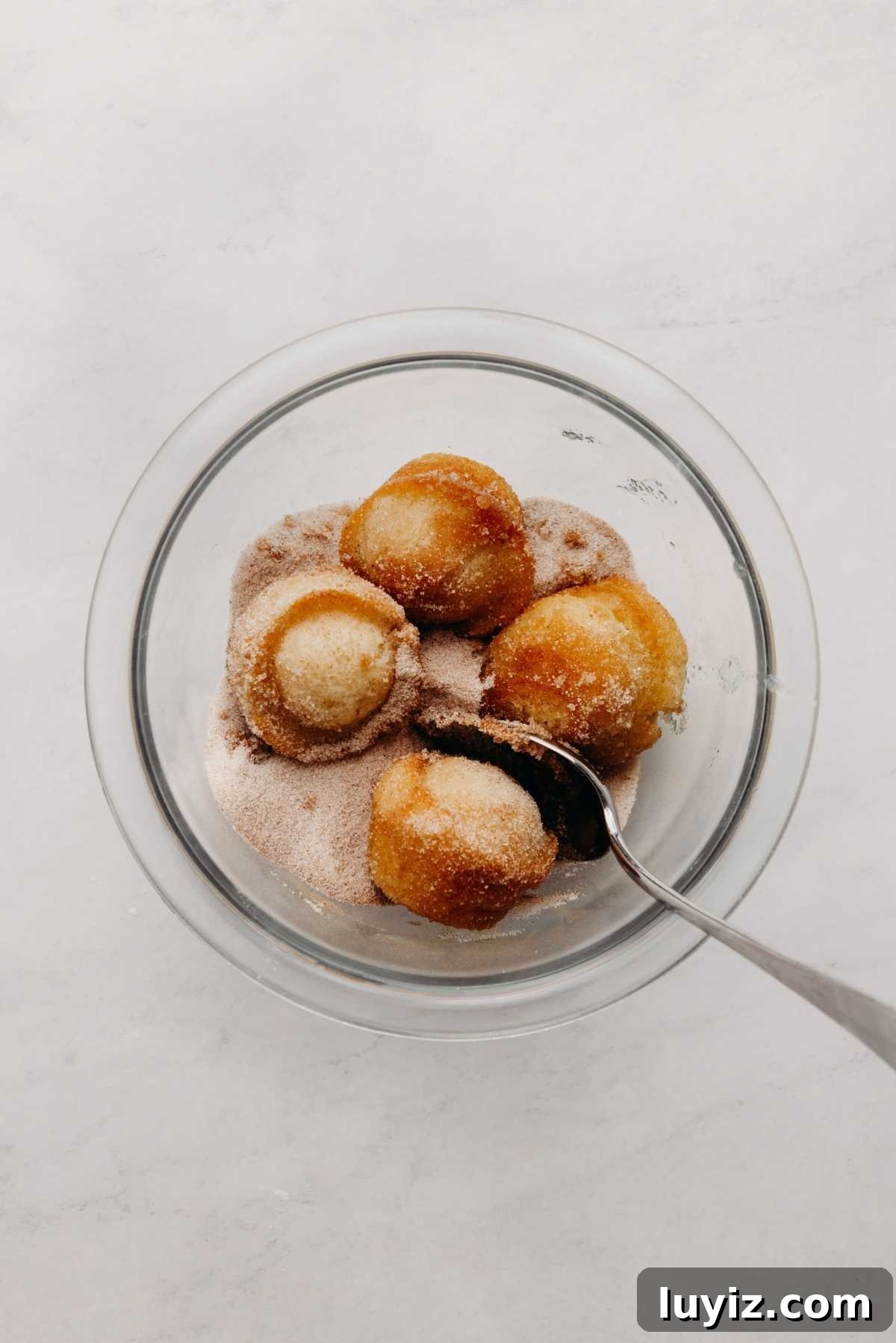 Mini French Breakfast Puffs: Airy, Jam-Filled Pastry Bites 13 Coating mini breakfast muffins in cinnamon sugar mix using a spoon in a small glass bowl on a white marbled background.