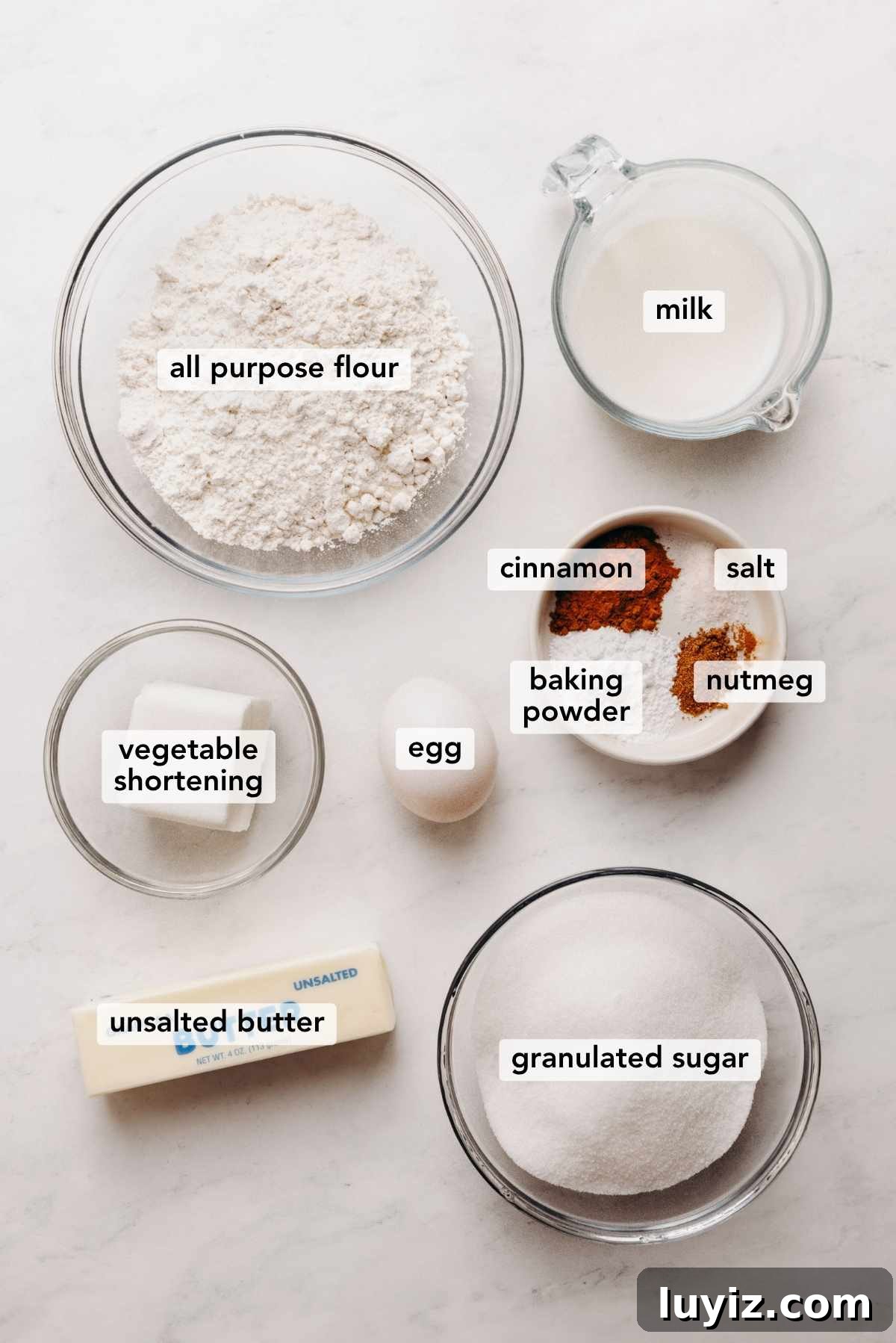 Mini French Breakfast Puffs: Airy, Jam-Filled Pastry Bites 5 Ingredients in various size bowls labeled as all purpose flour, milk, vegetable shortening, cinnamon, salt, baking powder, nutmeg, egg, unsalted butter, and granulated sugar on a white marbled background.