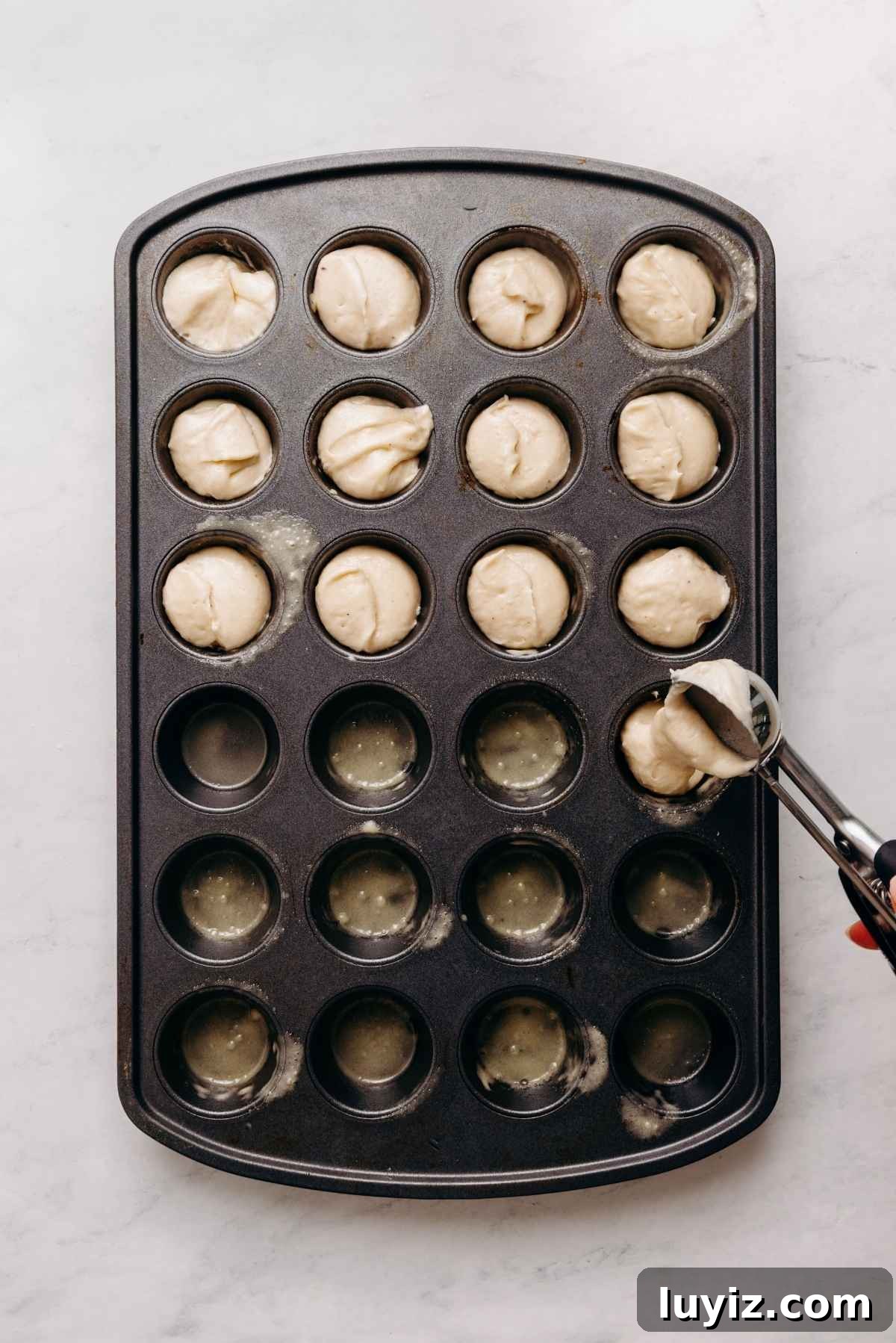 Mini French Breakfast Puffs: Airy, Jam-Filled Pastry Bites 10 Scooping breakfast muffin batter into coated mini muffin cavities on a white marbled background.