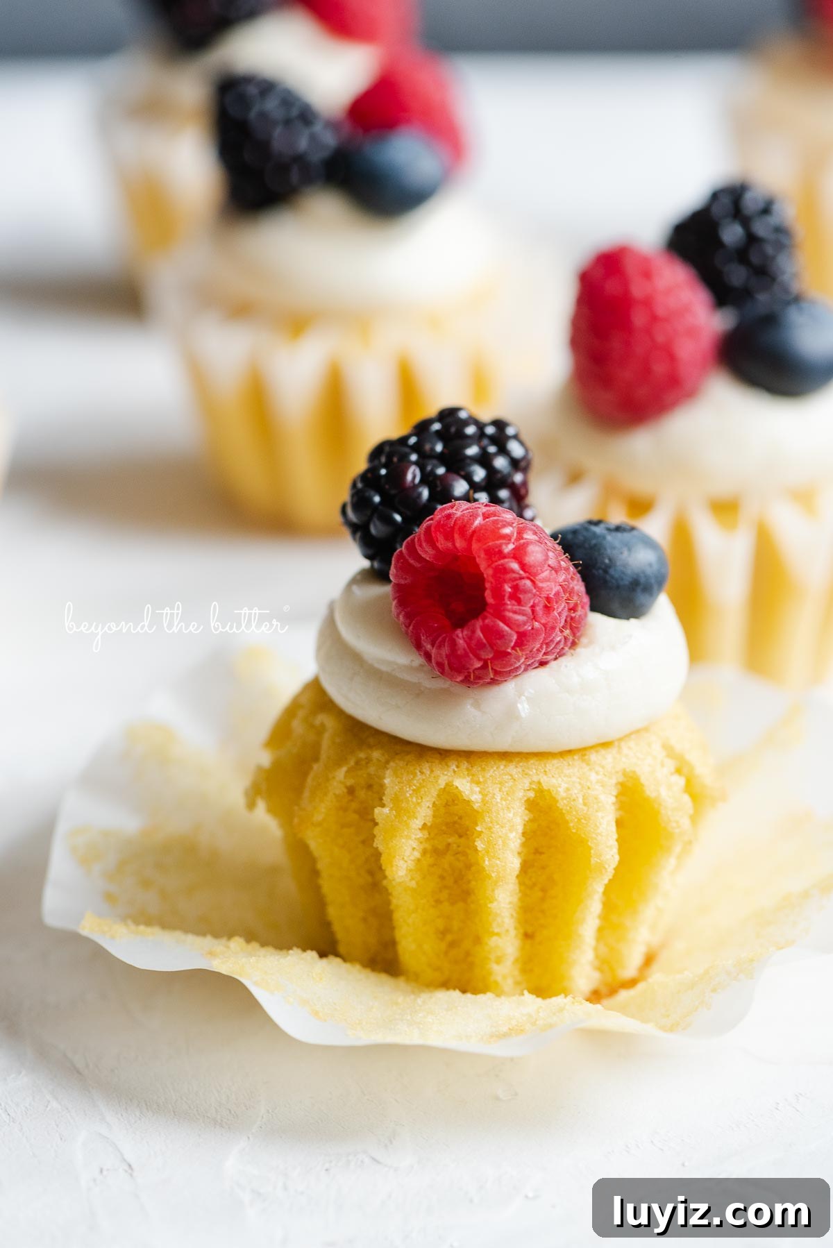 Mini Vanilla Cupcakes Recipe for Small Batches 3 Angled image of small batch vanilla cupakes topped with vanilla buttercream frosting and berries with front cupcake unwrapped.