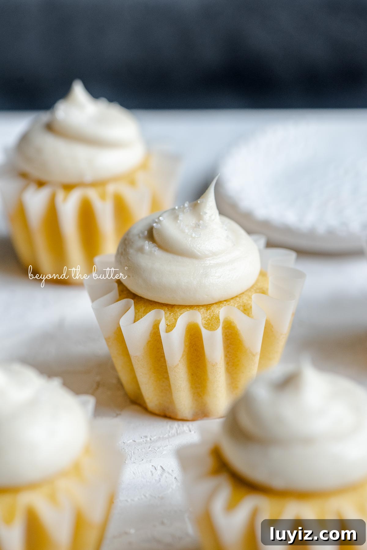 Mini Vanilla Cupcakes Recipe for Small Batches 4 Angled image of frosted small batch vanilla cupcakes sprinkled with sparkling sugar randomly placed on white and dark blue background next to a small shallow white bowl of sparkling sugar.