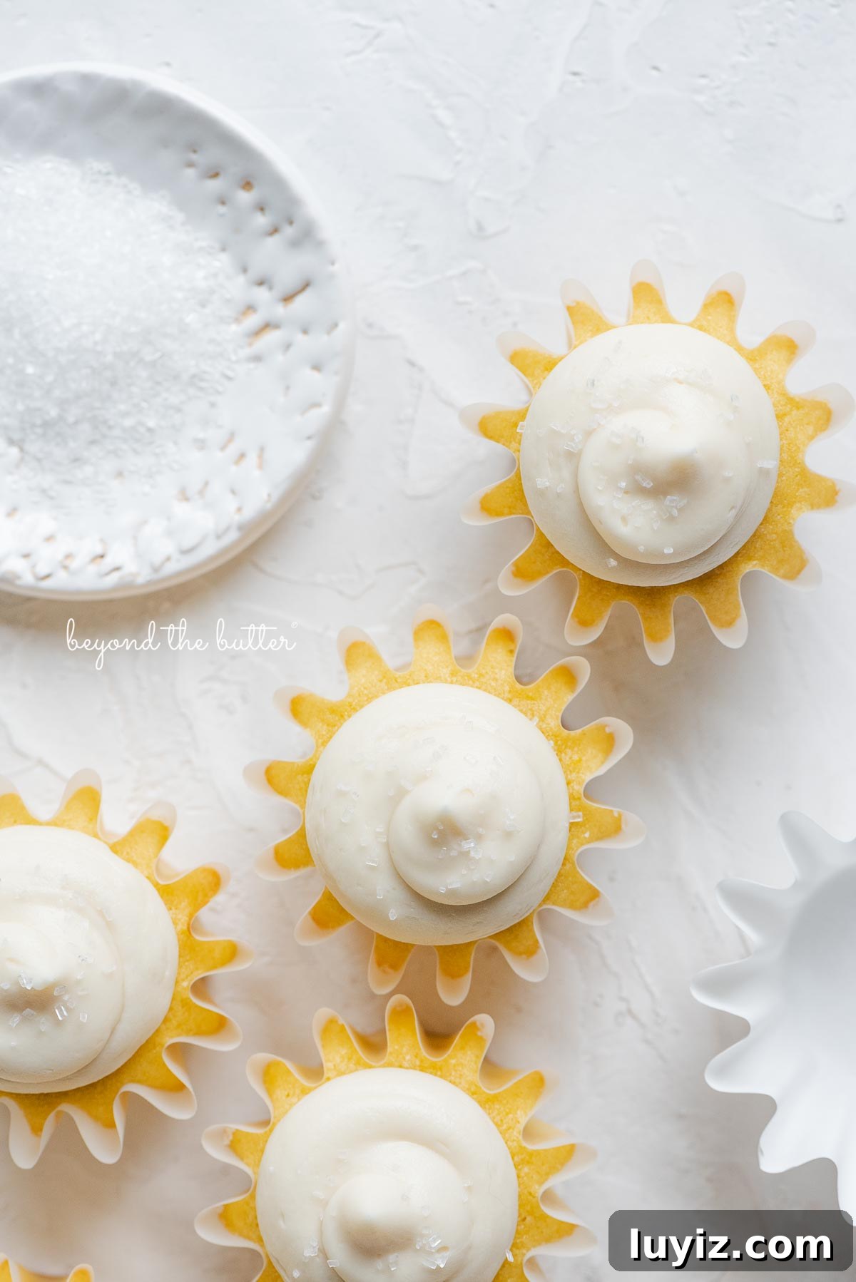 Mini Vanilla Cupcakes Recipe for Small Batches 6 Overhead image of frosted vanilla cupcakes sprinkled with sparkling sugar randomly placed on white background next to empty cupcake liner and small shallow bowl of white sparkling sugar.