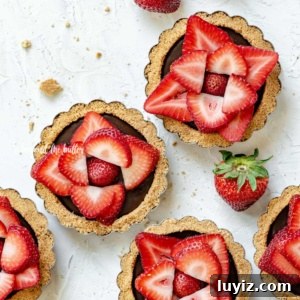 Mini Strawberry Nutella Tartlets: Bite-Sized Dessert Recipe 8 Overhead image of 6 mini strawberry nutella tarts on white stucco background with randomly placed strawberries around them.