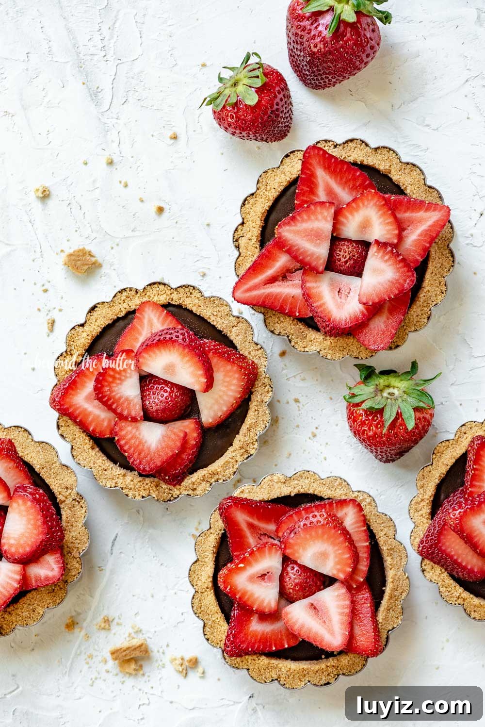 Mini Strawberry Nutella Tartlets: Bite-Sized Dessert Recipe 3 Overhead image of 6 mini strawberry nutella tarts on white stucco background with randomly placed strawberries around them | All Images © Beyond the Butter™