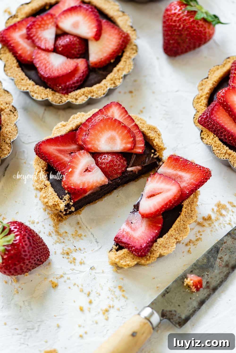 Mini Strawberry Nutella Tartlets: Bite-Sized Dessert Recipe 5 Angled image of mini strawberry nutella tarts on white stucco background with one sliced in half and opened | All Images © Beyond the Butter™
