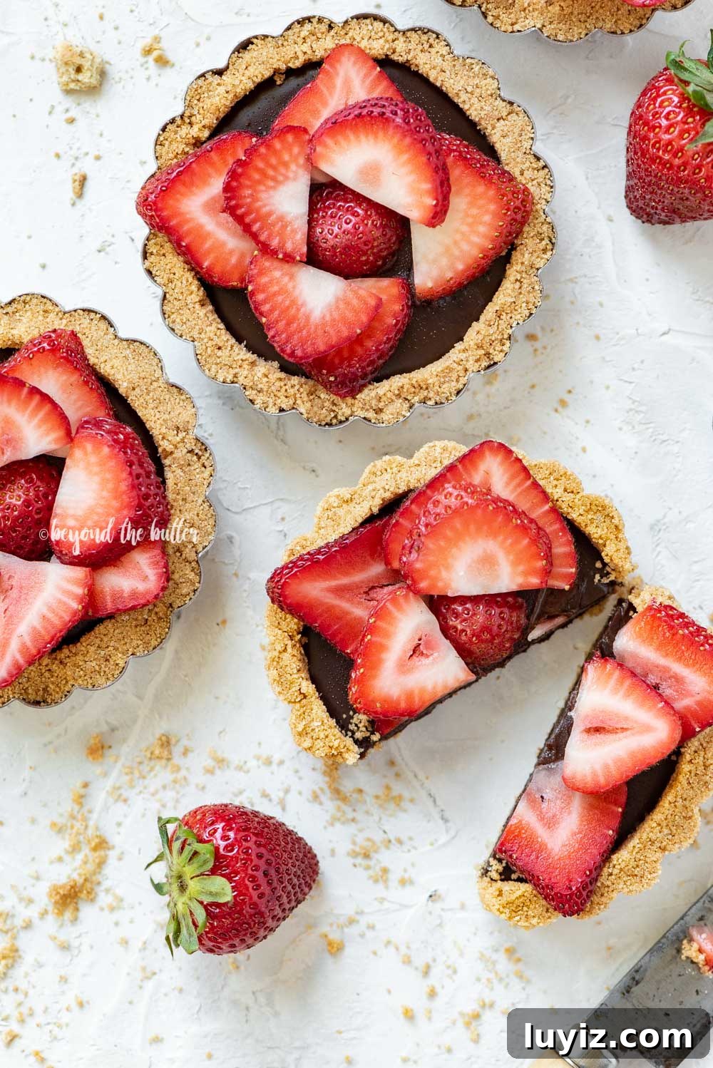 Mini Strawberry Nutella Tartlets: Bite-Sized Dessert Recipe 7 Overhead image of mini strawberry nutella tarts on white stucco background with one sliced in half and opened | All Images © Beyond the Butter™