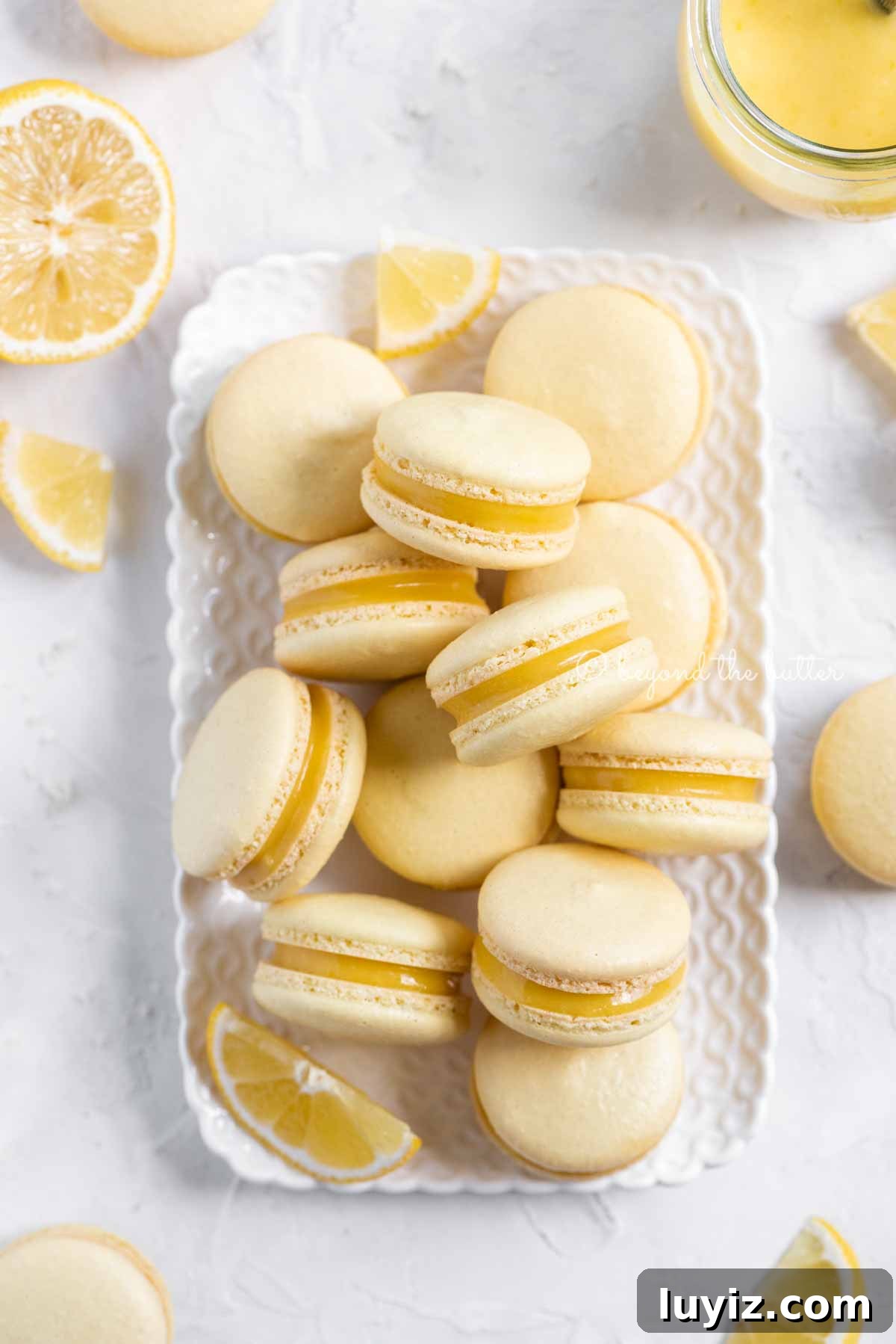 Zesty Lemon Bar Macarons: Tangy Citrus Sandwich Cookies 4 Overhead image of lemon bar macarons on a white dessert tray with lemon wedges | All Images © Beyond the Butter™