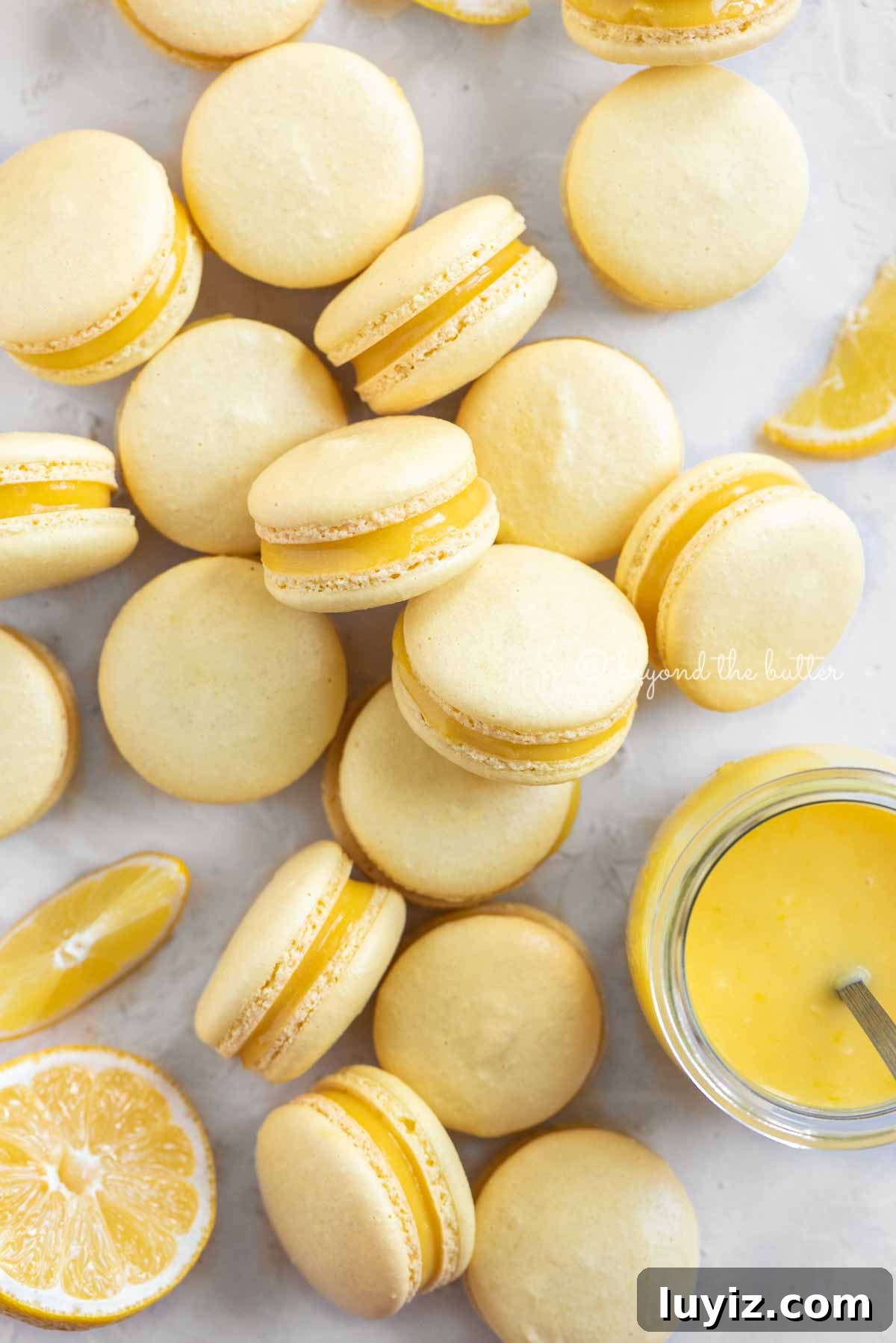 Zesty Lemon Bar Macarons: Tangy Citrus Sandwich Cookies 5 Overhead image of lemon bar macarons randomly placed around a small jar of lemon curd filling | All Images © Beyond the Butter™