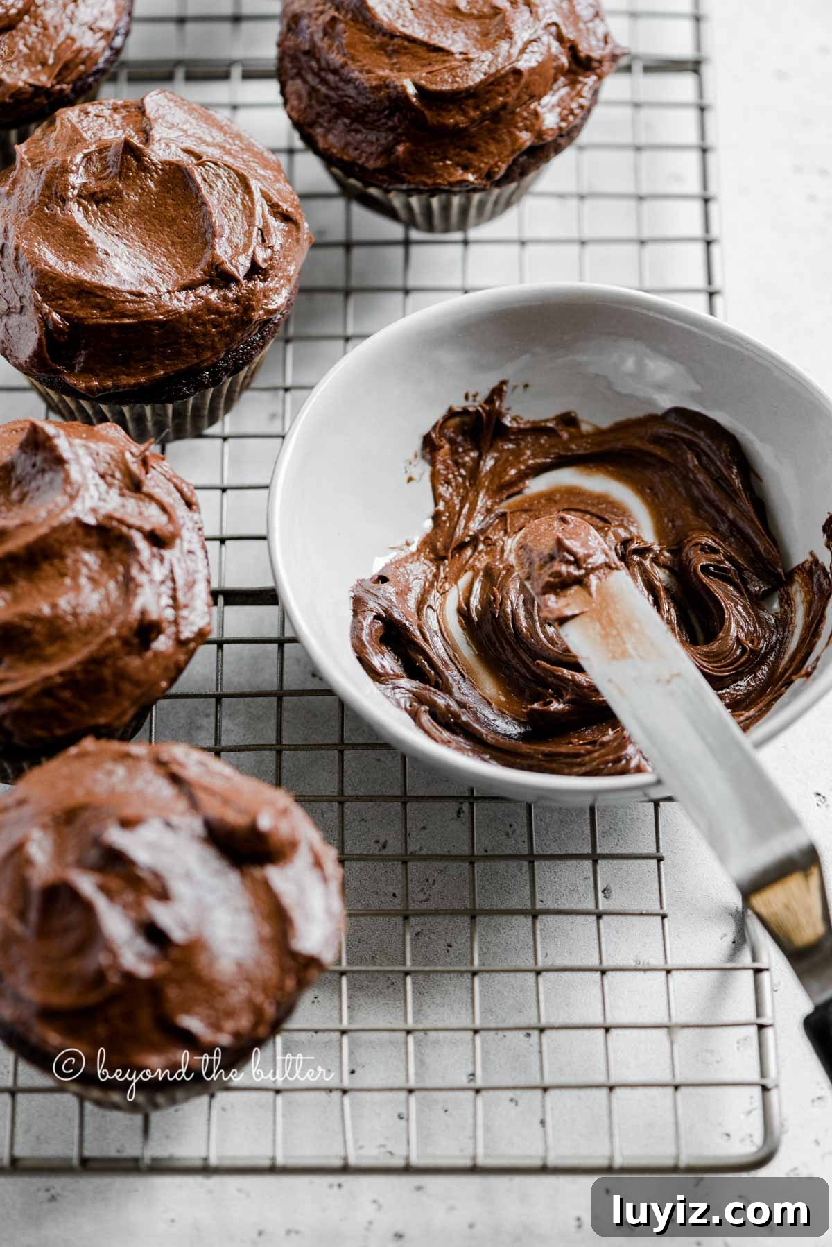Small-Batch Chocolate Cupcakes Recipe for 12 Moist Treats 3 Angled image of chocolate cupcakes on cooling rack with small bowl of chocolate frosting and offset spatula | All Images © Beyond the Butter™