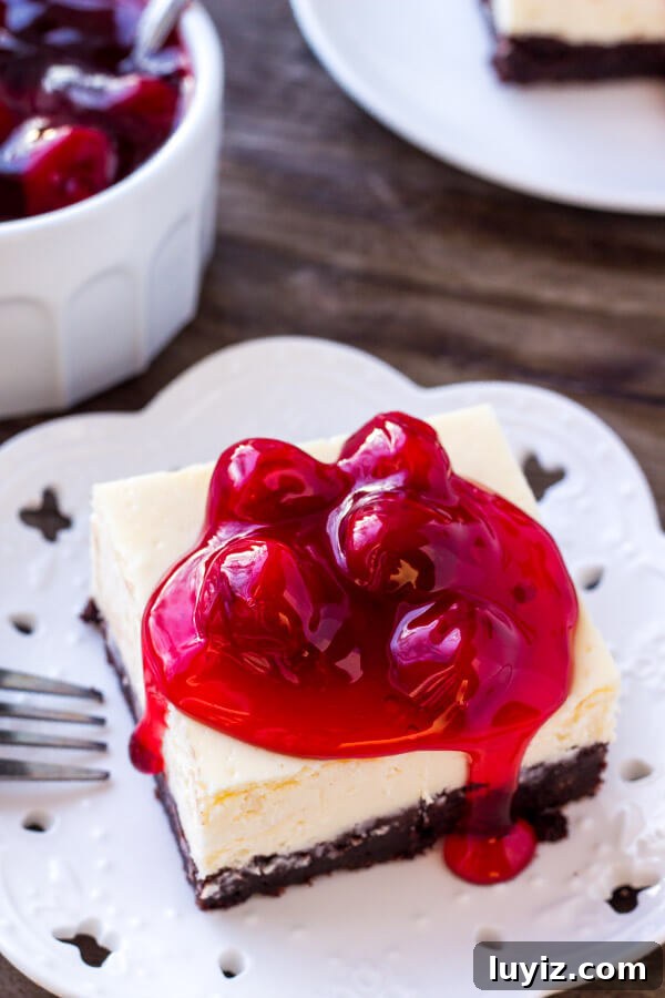 Overhead view of a slice of brownie cheesecake bars topped with cherries. With a bowl of cherry topping in the background.