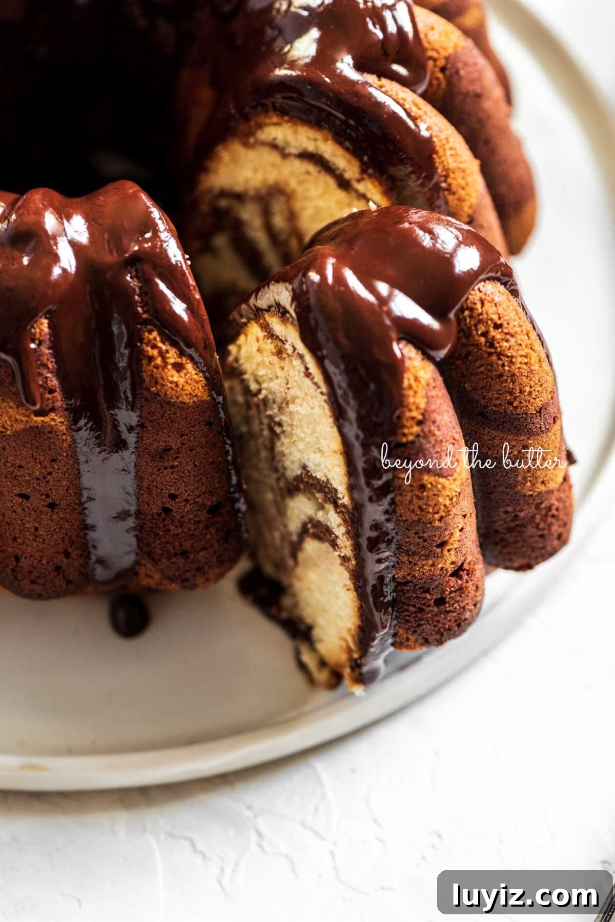 Decadent Marble Bundt Cake Recipe with Swirled Chocolate-Vanilla 2 Chocolate glazed marble bundt cake with one slice partially removed on a white ceramic dessert plate and white background.