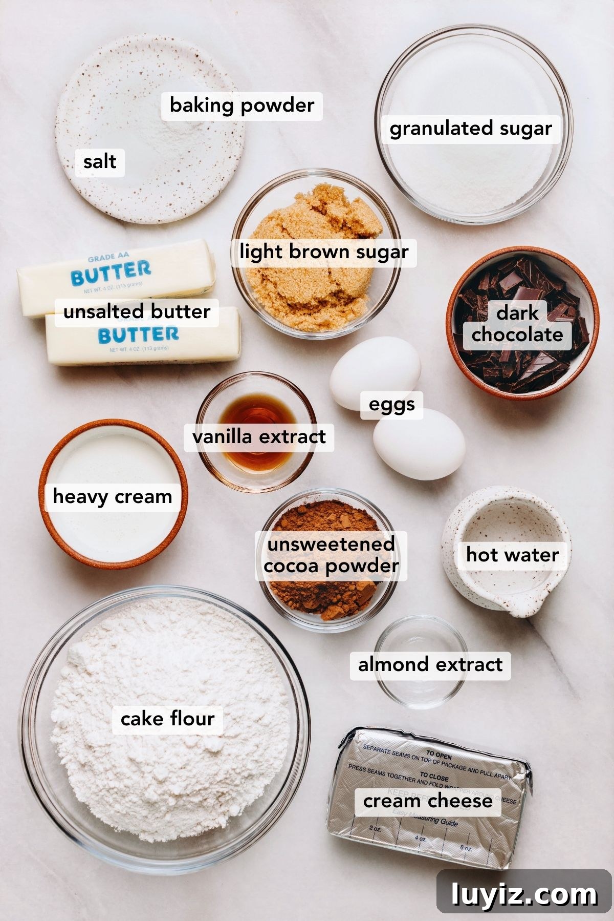 Decadent Marble Bundt Cake Recipe with Swirled Chocolate-Vanilla 4 Ingredients in various size bowls and plates labeled as salt, baking powder, granulated sugar, light brown sugar, unsalted butter, heavy cream, vanilla extract, almond extract, cream cheese, hot water, unsweetened cocoa powder, cake flour, dark chocolate, and eggs on a white marbled background.