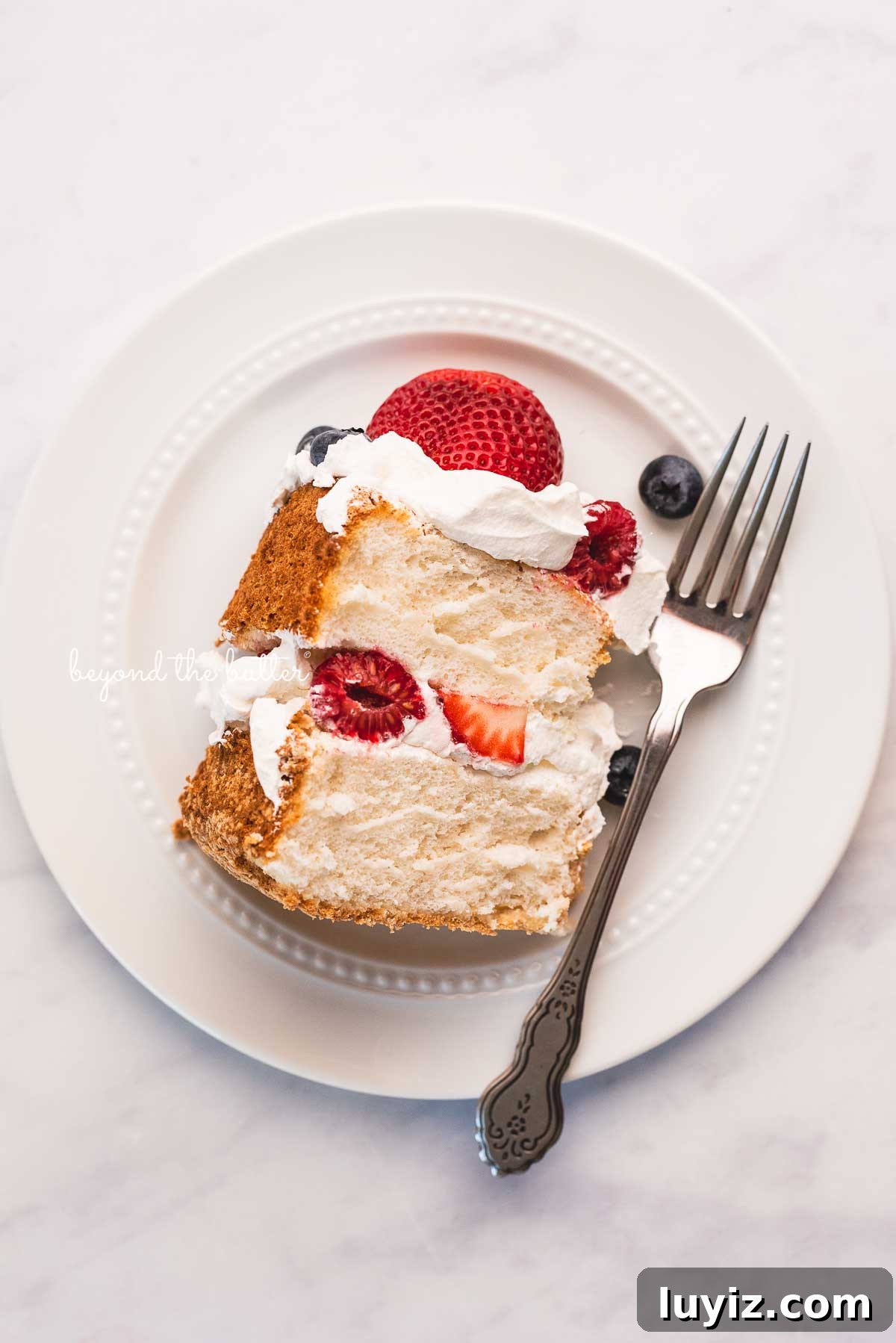 Light and Fluffy Homemade Angel Food Cake Recipe 3 Slice of homemade angel food cake with berries on white dessert plate and white marbled background
