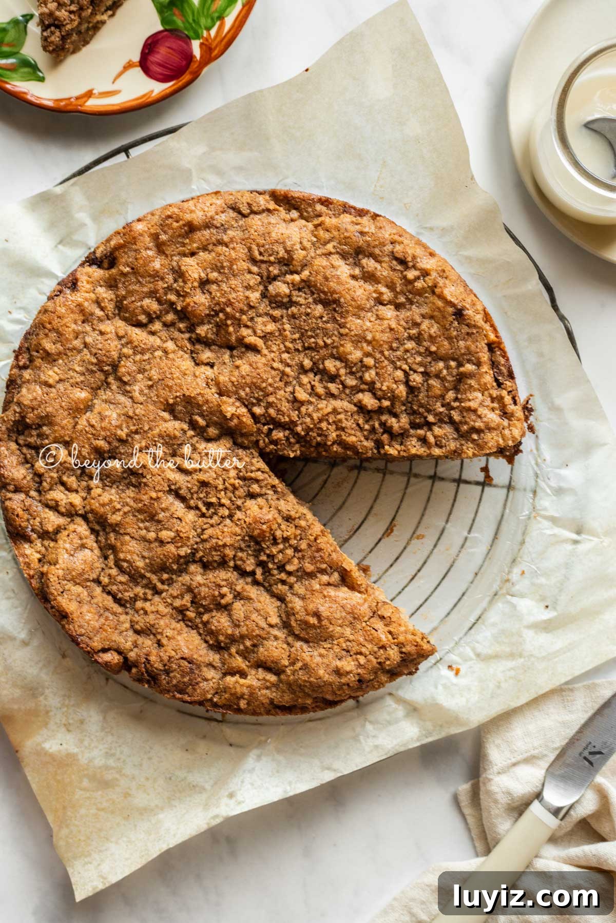 Apple Cinnamon Coffee Cake Recipe for Moist, Tender Crumbs 8 Apple coffee cake on a parchment paper lined wire cooling rack with a slice removed and a simple vanilla glaze, dessert plates, and a knife to the side | All Images © Beyond the Butter™